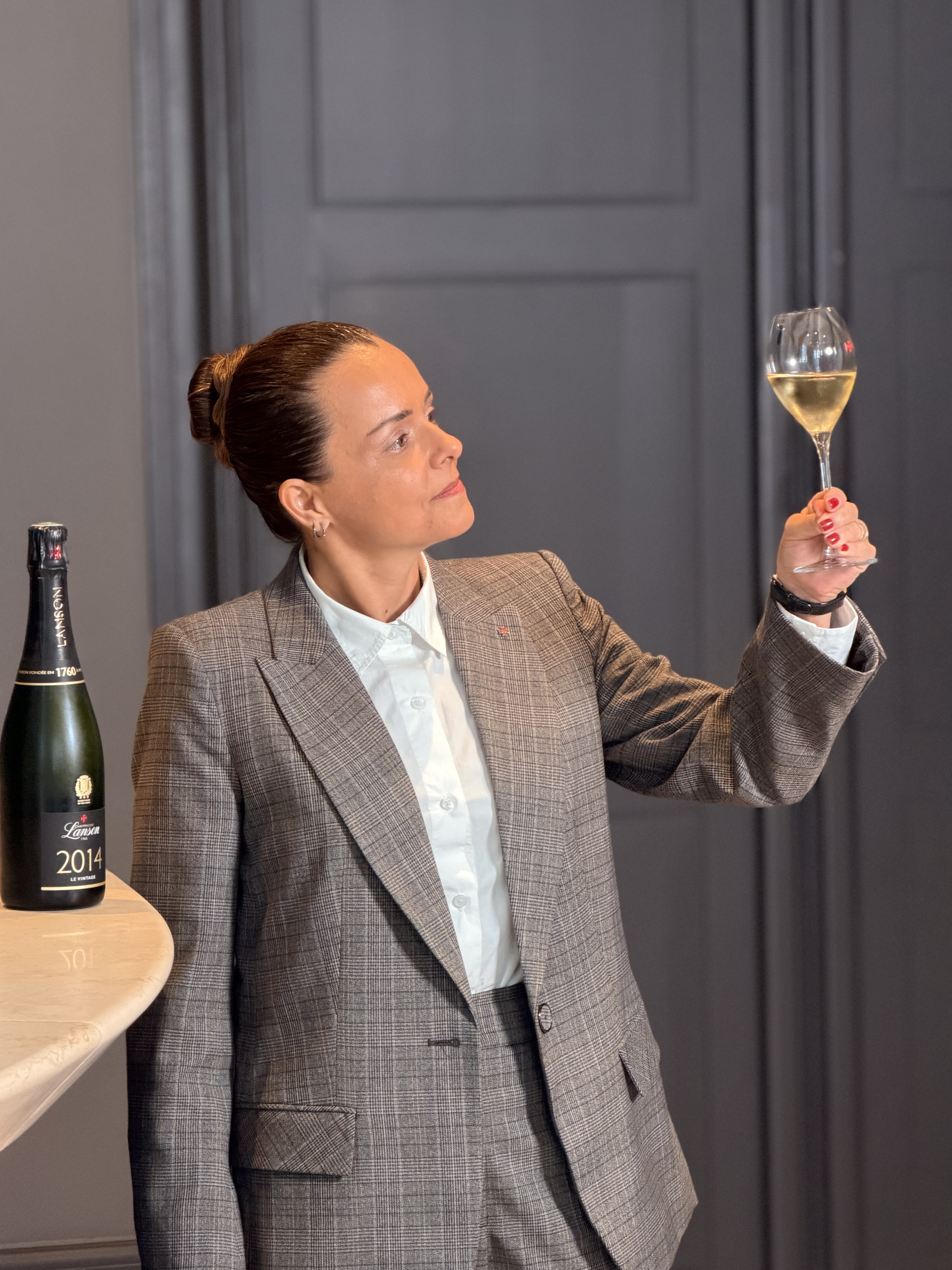 Mulher de cabelo preso, camisa branca e terno xadrez marrom, segurando uma ta&ccedil;a de vinho branco e observando o l&iacute;quido. Ao lado, uma garrafa de champanhe verde com r&oacute;tulo Lanson 2014 sobre uma mesa clara. O fundo &eacute; cinza escuro