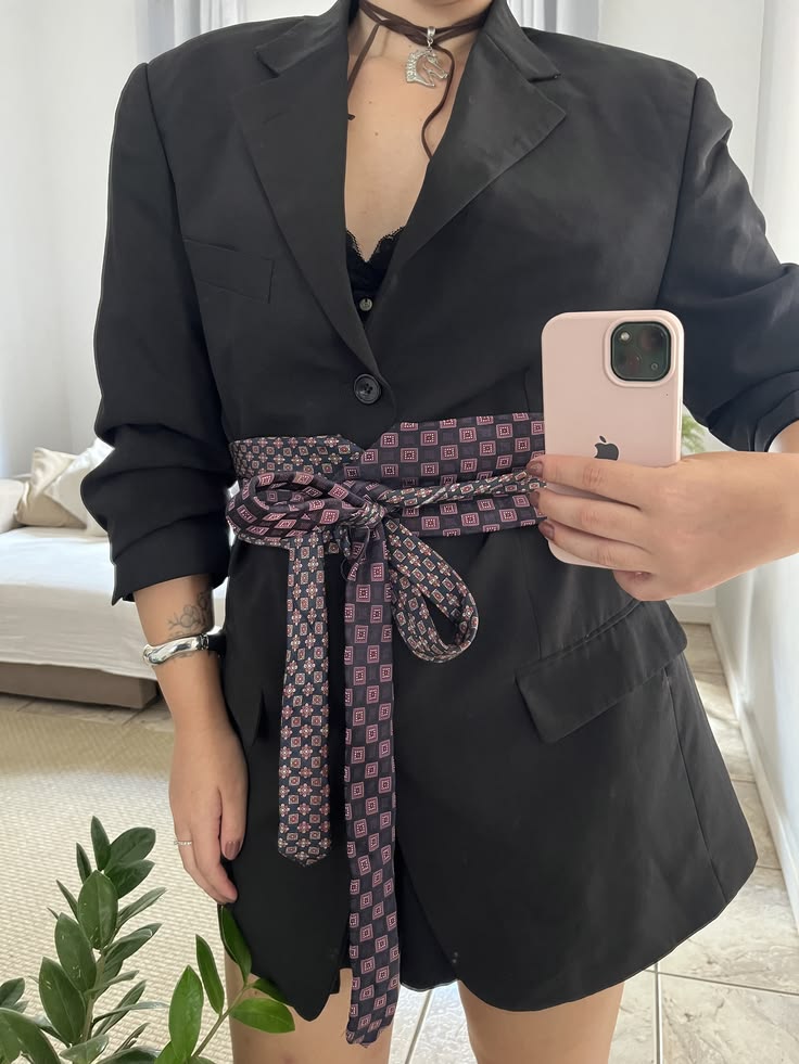 Pessoa de p&eacute;, sem rosto vis&iacute;vel, vestindo um blazer preto com um cinto de tecido estampado em tons de roxo e preto. Ela segura um celular rosa claro na m&atilde;o direita, com uma pulseira prateada e anel na m&atilde;o esquerda. Ao fundo, um sof&aacute; claro e uma planta verde.