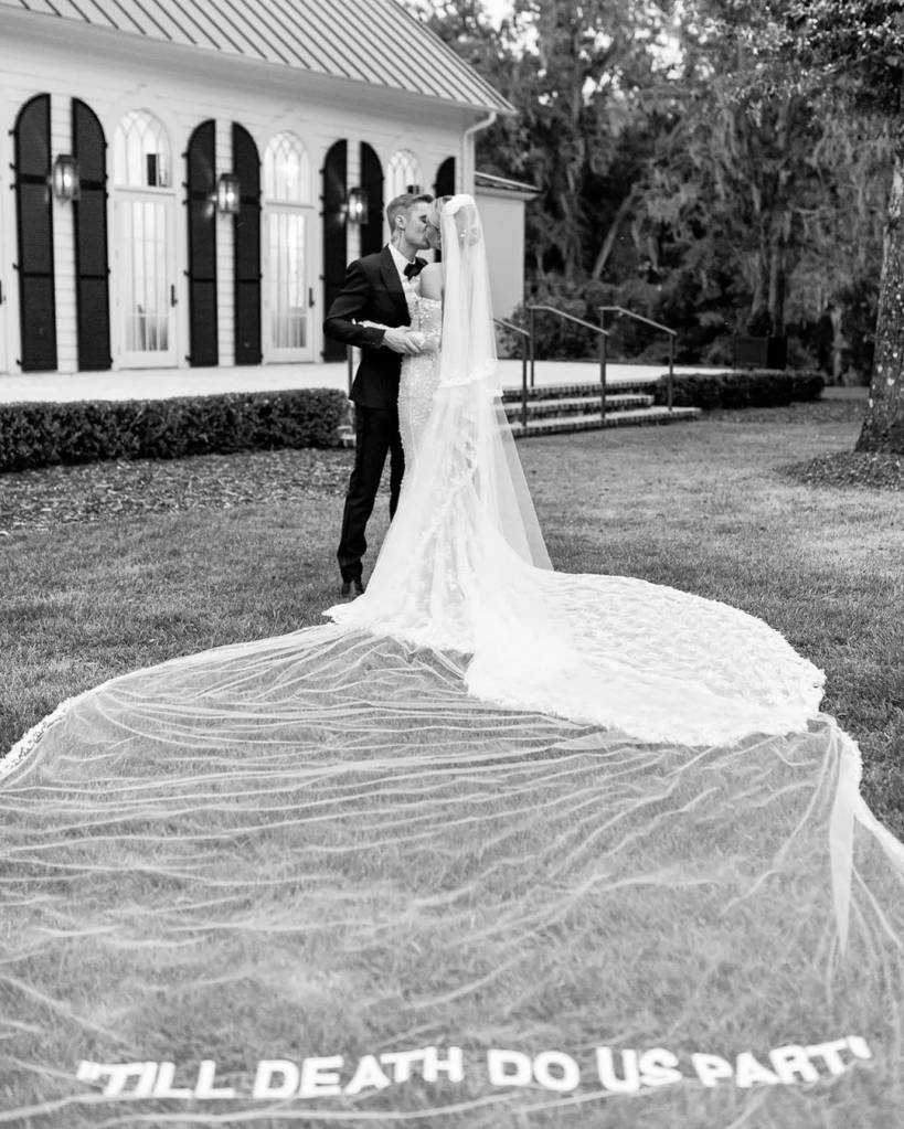 Casal de noivos se beijando em preto e branco, com o v&eacute;u da noiva estendido no gramado e a frase TILL DEATH DO US PART escrita nele. Ao fundo, uma casa branca com janelas escuras e &aacute;rvores