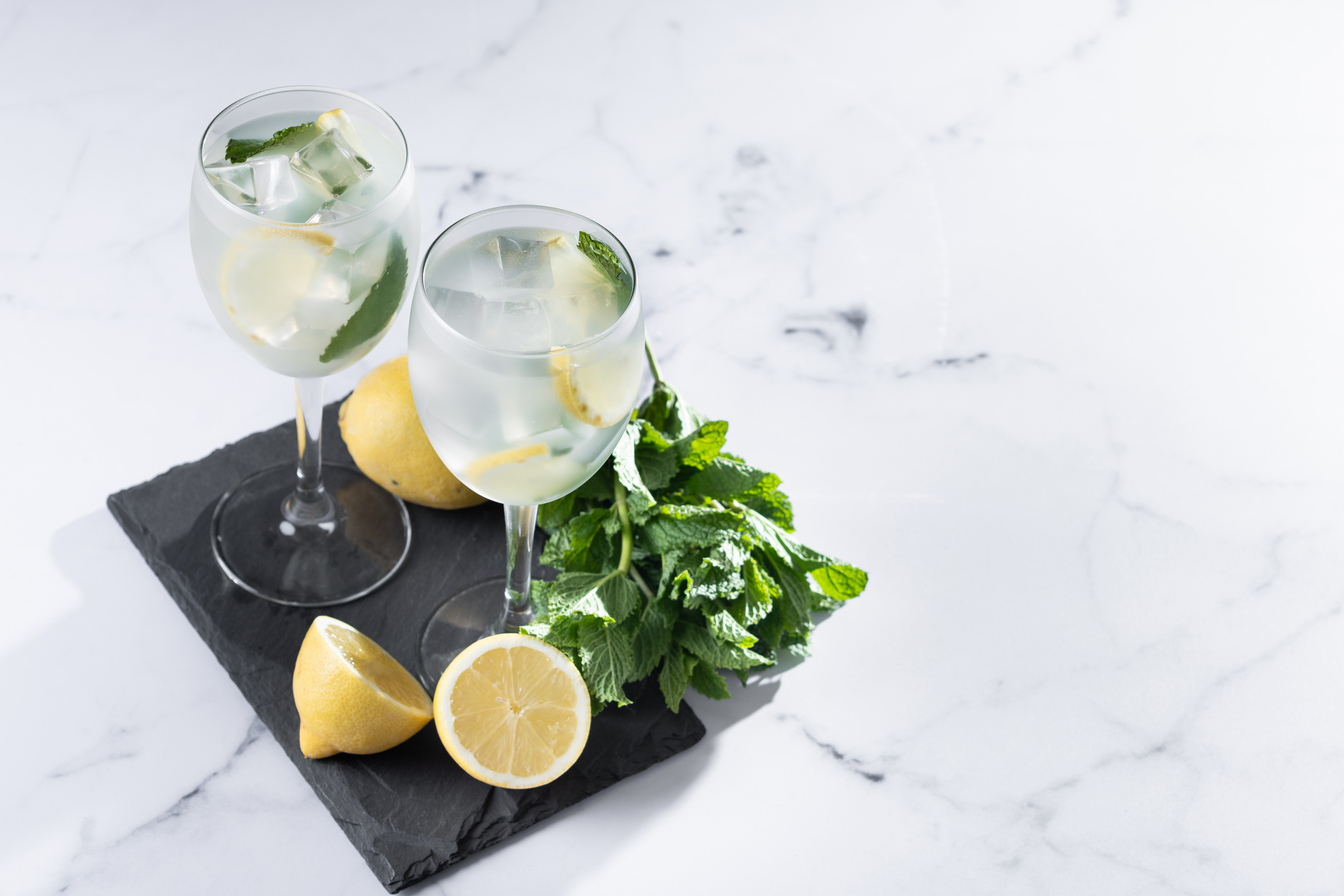 Dois copos de gin tônica com gelo, fatias de limão e folhas de hortelã, sobre uma tábua preta de ardósia. Ao lado, um limão inteiro e outro cortado ao meio, além de um ramo de hortelã fresca. Tudo disposto sobre uma superfície de mármore branco com veios cinzas, iluminada por luz natural