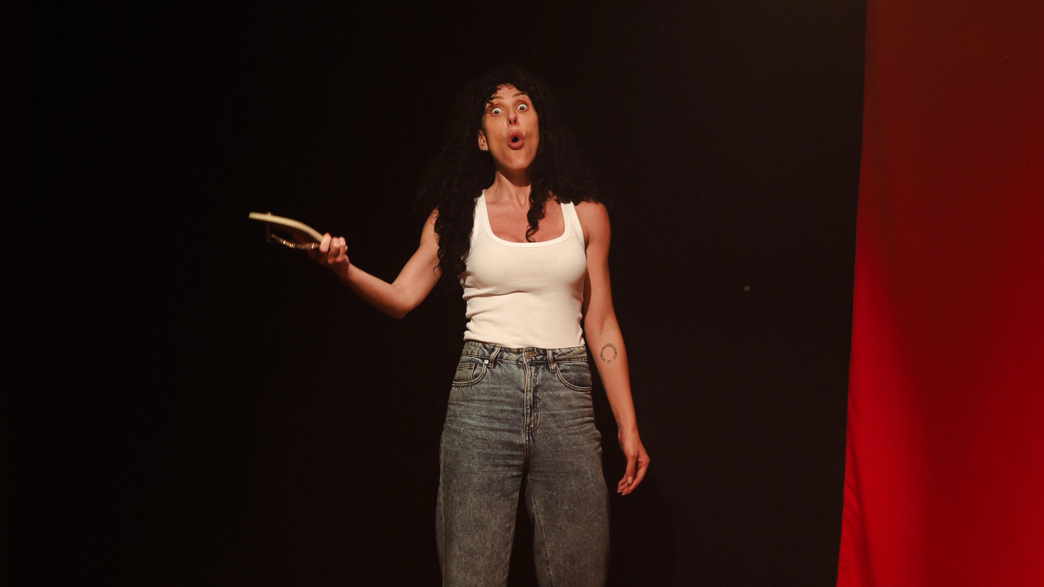 Mulher de cabelos escuros e cacheados, vestindo regata branca e cal&ccedil;a jeans, com express&atilde;o de surpresa e boca aberta, segurando um espelho oval na m&atilde;o esquerda, em um palco com fundo preto e uma faixa vertical vermelha &agrave; direita