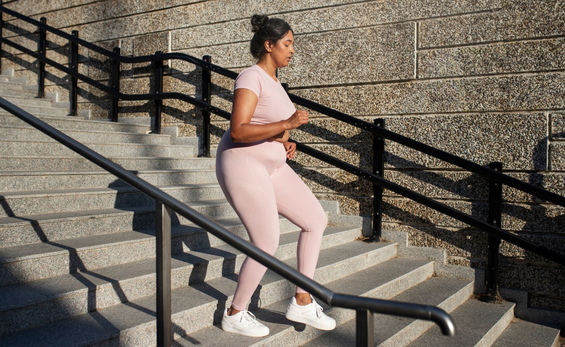Mulher de pele morena e cabelo preso, vestindo top e calça legging rosa claro, sobe uma escadaria de granito cinza com corrimãos pretos, em um dia ensolarado