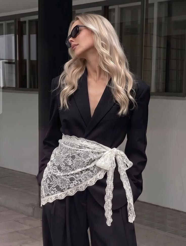 Mulher loira de &oacute;culos escuros, blazer e cal&ccedil;a pretos, com uma faixa de renda branca amarrada na cintura, posando em frente a um pr&eacute;dio com janelas escuras