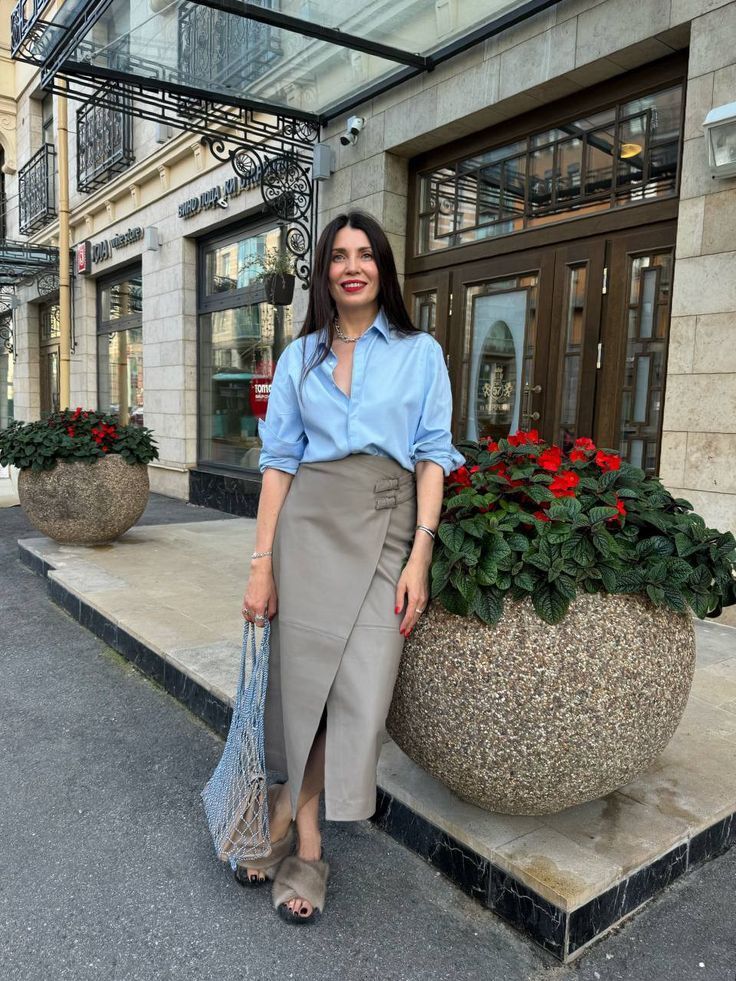 Mulher de cabelos longos e escuros, camisa azul clara e saia midi cinza-esverdeada, sorrindo em frente a uma loja com vasos de flores vermelhas. Ela segura uma bolsa de rede azul e usa sand&aacute;lias de pelo.
