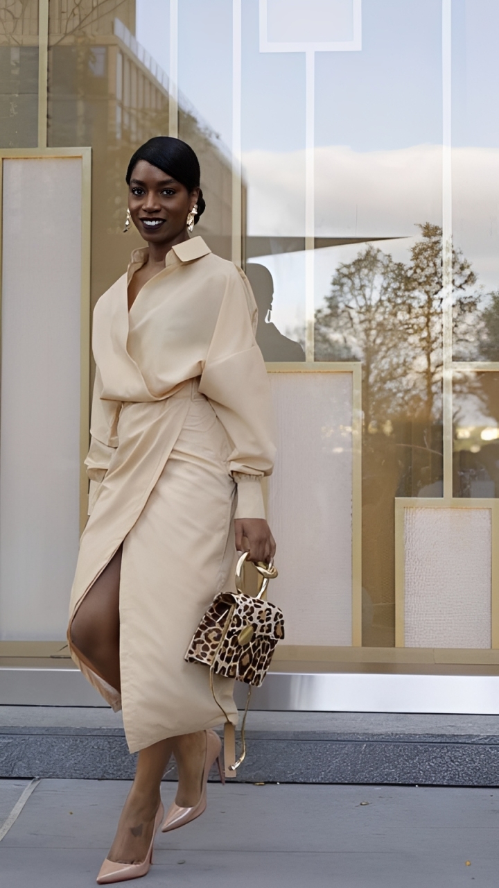 Mulher negra sorrindo, cabelo preso, usando brincos dourados e um conjunto bege de blusa transpassada e saia com fenda. Ela segura uma bolsa de oncinha com al&ccedil;a dourada e cal&ccedil;a sapatos de salto nude, posando em frente a uma vitrine com reflexos de &aacute;rvores