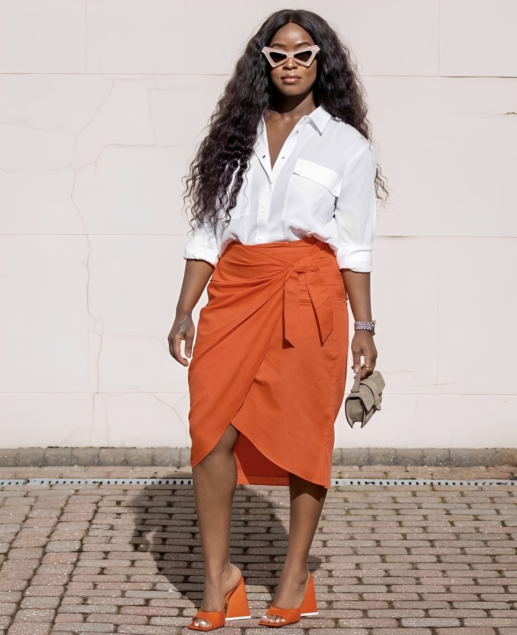 Mulher negra de cabelos longos e ondulados, usando &oacute;culos de sol rosa-claro, camisa branca de bot&otilde;es, saia envelope laranja com amarra&ccedil;&atilde;o na cintura, sand&aacute;lias de salto laranja e bolsa pequena bege, posando em frente a uma parede clara e um ch&atilde;o de tijolos