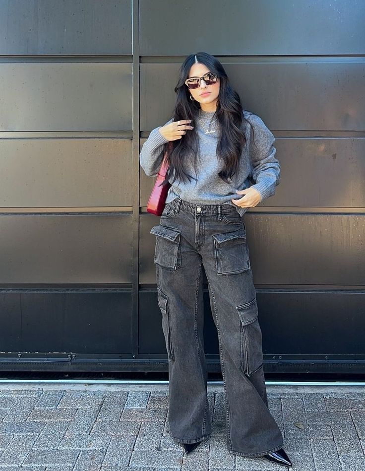 Mulher jovem com cabelos longos e escuros, &oacute;culos de sol, su&eacute;ter cinza e cal&ccedil;a cargo jeans escura, posando em frente a uma parede preta e um ch&atilde;o de paralelep&iacute;pedos