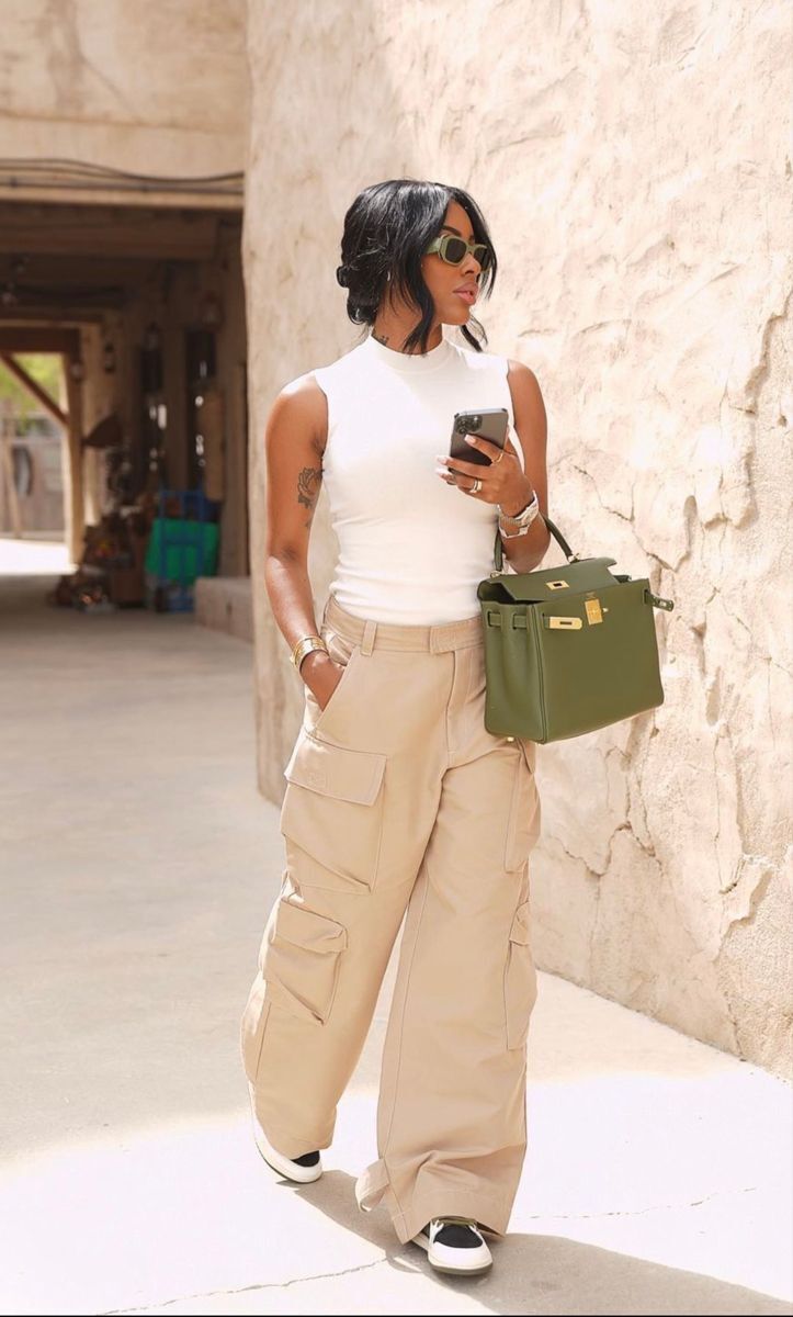 Mulher negra com cabelo escuro preso, &oacute;culos de sol verde-oliva, regata branca e cal&ccedil;a cargo bege larga, segurando um celular e uma bolsa verde-militar, em p&eacute; ao lado de uma parede de pedra clara