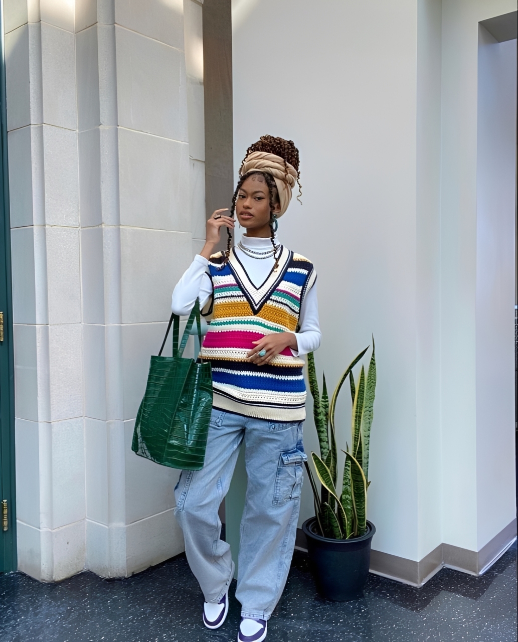 Mulher jovem com turbante bege e cabelo cacheado, vestindo su&eacute;ter de tric&ocirc; listrado colorido sobre blusa branca, cal&ccedil;a cargo jeans clara e t&ecirc;nis branco e roxo, segurando bolsa verde texturizada. Ela posa em p&eacute;, olhando para frente, ao lado de uma planta em vaso preto e uma parede branca com detalhes arquitet&ocirc;nicos