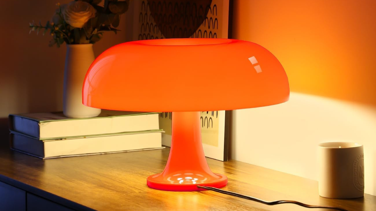 Uma luminária de mesa laranja brilhante com base e cúpula em formato de cogumelo, acesa em um ambiente com luz quente. À esquerda, um vaso branco com flores claras e dois livros empilhados. À direita, uma vela aromática em pote branco. O fundo é uma parede em tom laranja claro.