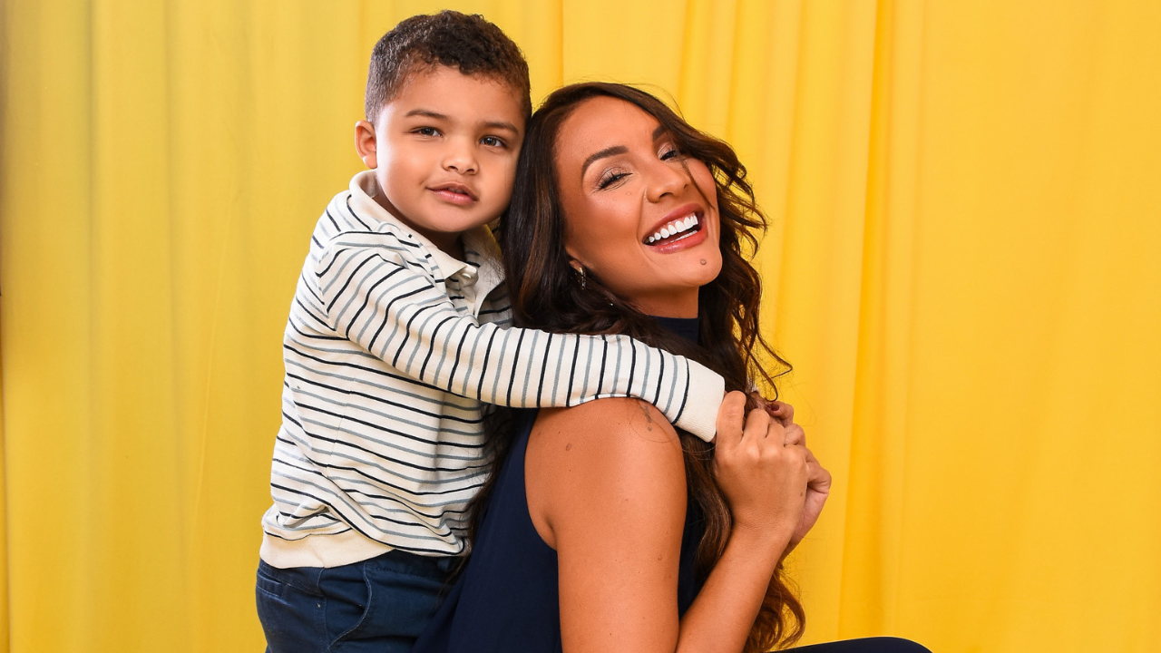 Mulher de cabelos longos e escuros, sorrindo com a boca aberta, abraçada por um menino de camisa listrada, ambos em frente a um fundo amarelo