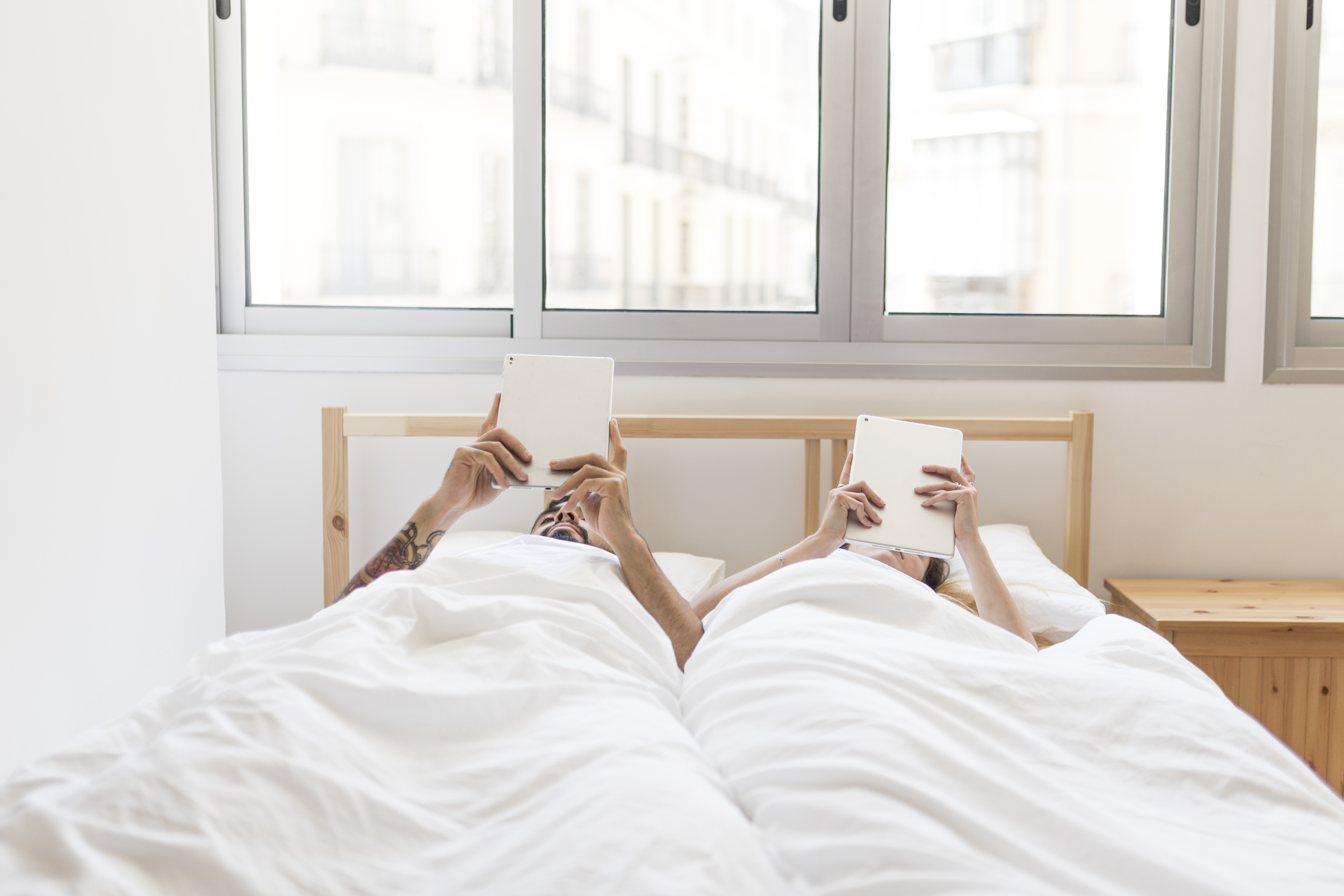 Casal deitado na cama, coberto por edredons brancos, cada um segurando um tablet branco acima da cabe&ccedil;a, com janelas ao fundo
