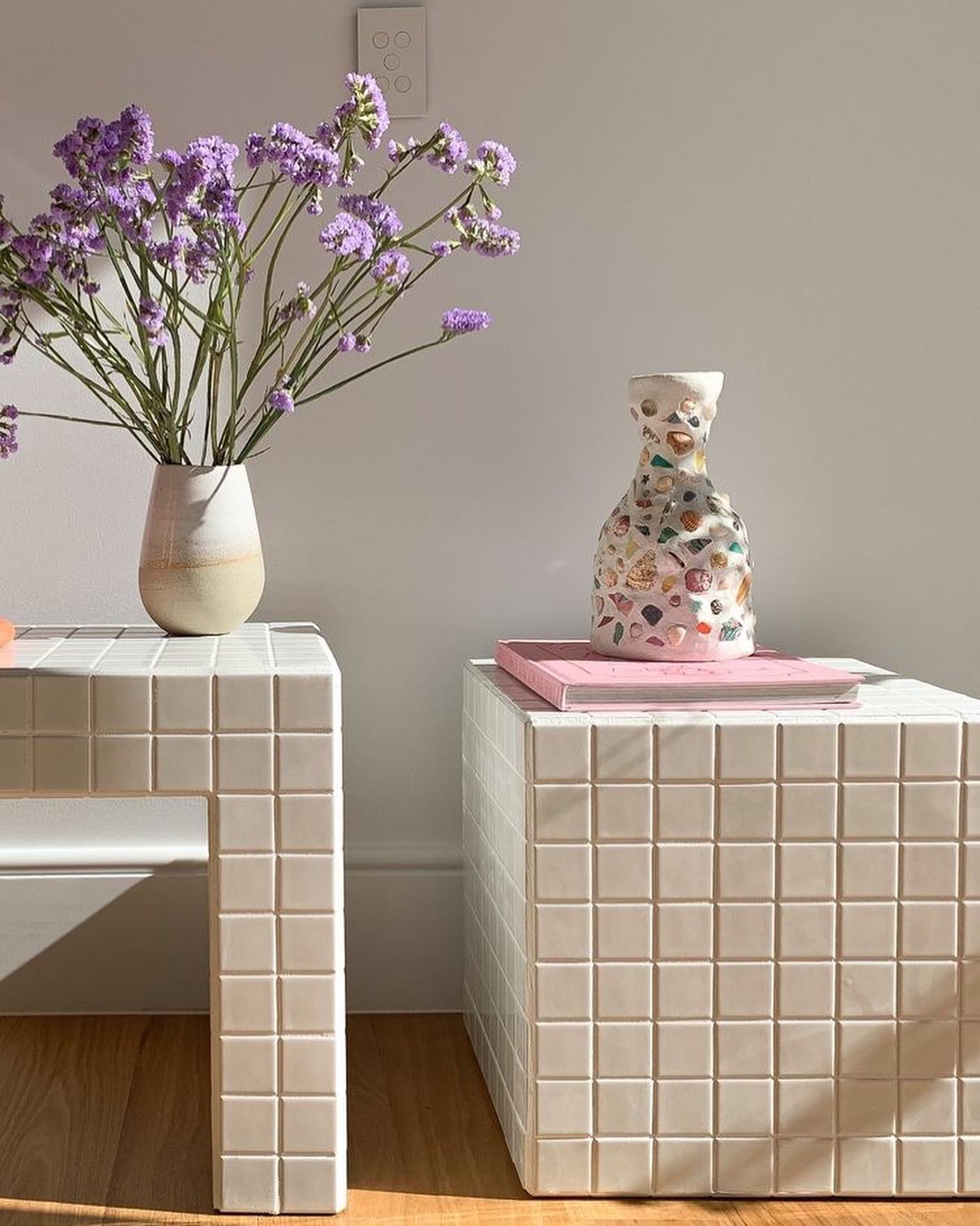 Duas mesas de cabeceira brancas com azulejos quadrados, uma em formato de U e outra c&uacute;bica. Na mesa em U, um vaso bege e branco com flores roxas. Na mesa c&uacute;bica, um vaso decorado com conchas e um livro rosa. O ambiente tem paredes brancas e piso de madeira, com luz solar entrando pela direita