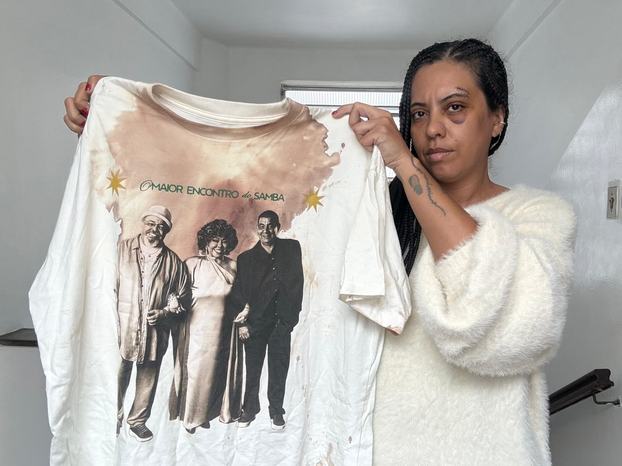 Mulher de tranças com olho roxo e corte na testa segura uma camiseta branca suja de sangue, estampada com três sambistas e a frase O MAIOR ENCONTRO do SAMBA