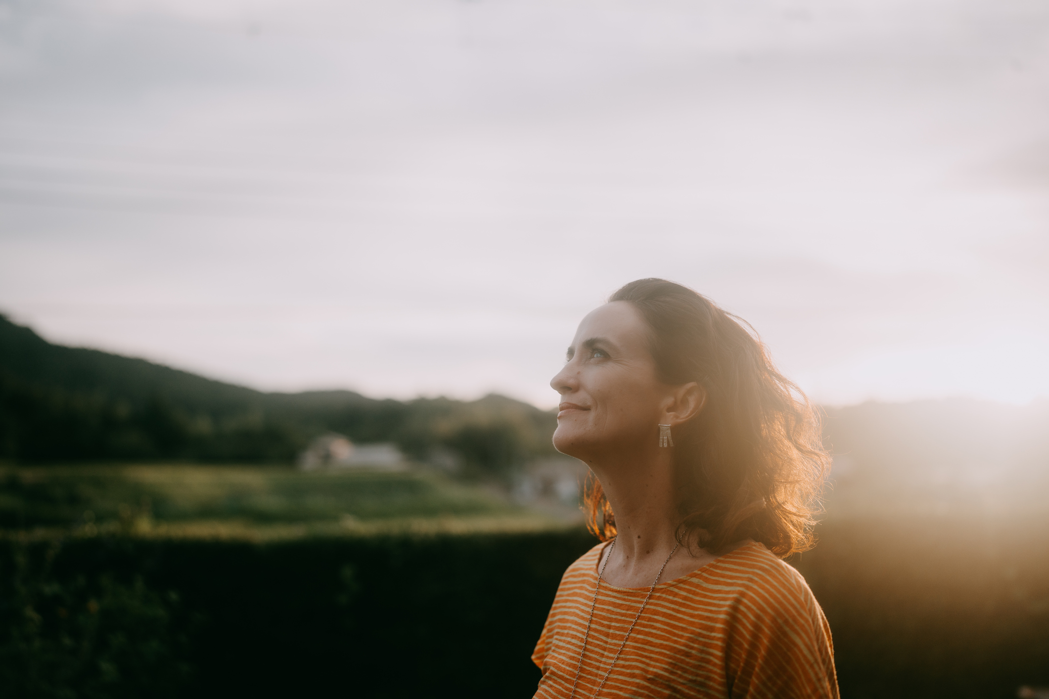 Mulher de cabelos castanhos e ondulados, vestindo camiseta laranja listrada, sorri olhando para cima, com o sol poente iluminando seu rosto e cabelo. Ao fundo, montanhas e casas embaçadas, com um céu claro e ensolarado