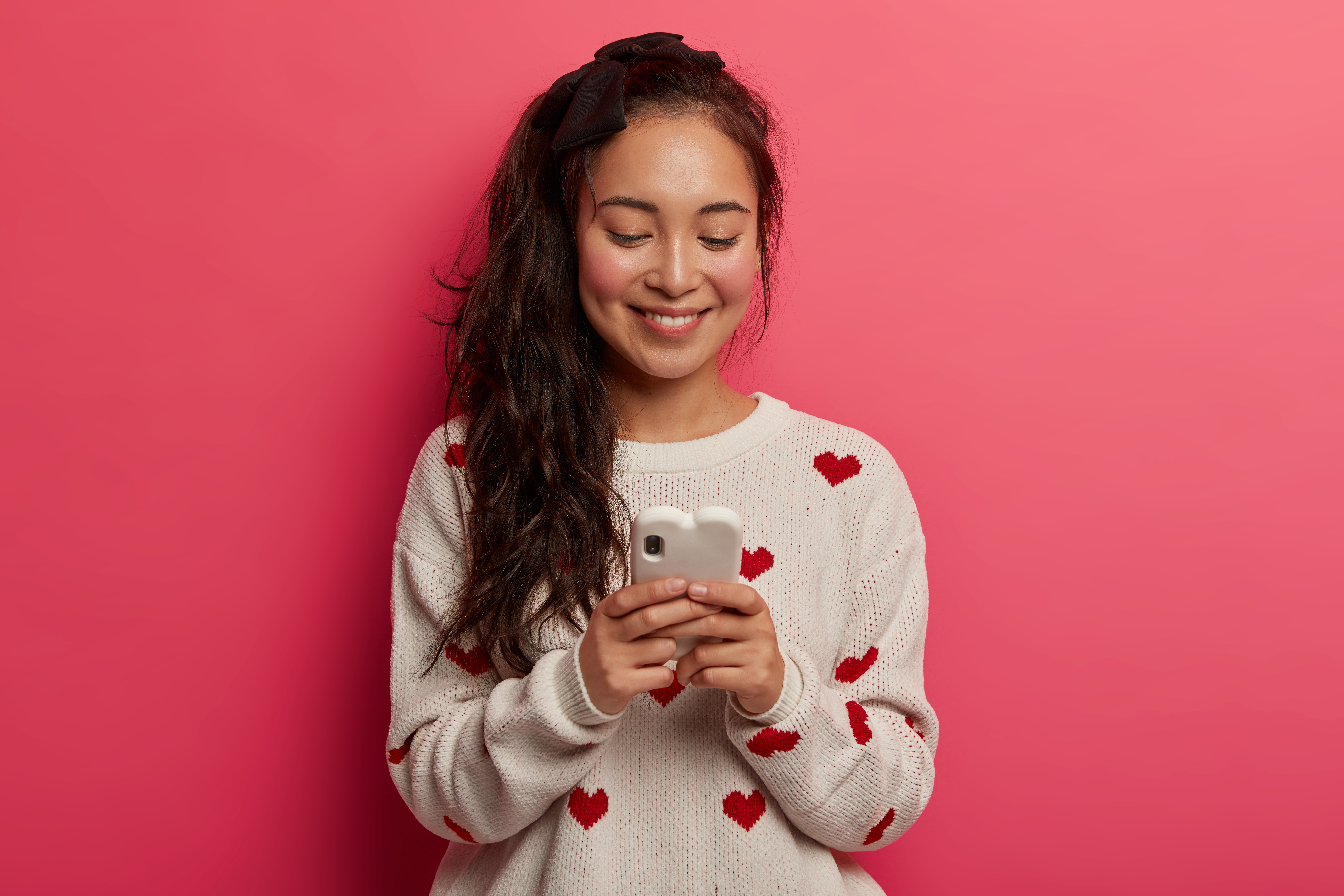 Jovem mulher asi&aacute;tica sorridente, com cabelo escuro e la&ccedil;o preto, usando um su&eacute;ter branco com cora&ccedil;&otilde;es vermelhos, olhando para o celular com uma capa em formato de cora&ccedil;&atilde;o, em um fundo rosa