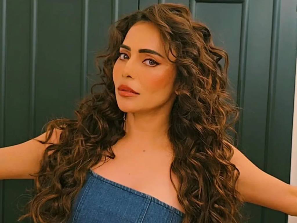 Mulher de pele clara, cabelos castanhos longos e cacheados, maquiagem marcante nos olhos e l&aacute;bios, vestindo um top jeans tomara que caia, posa com os bra&ccedil;os abertos contra uma parede verde escura