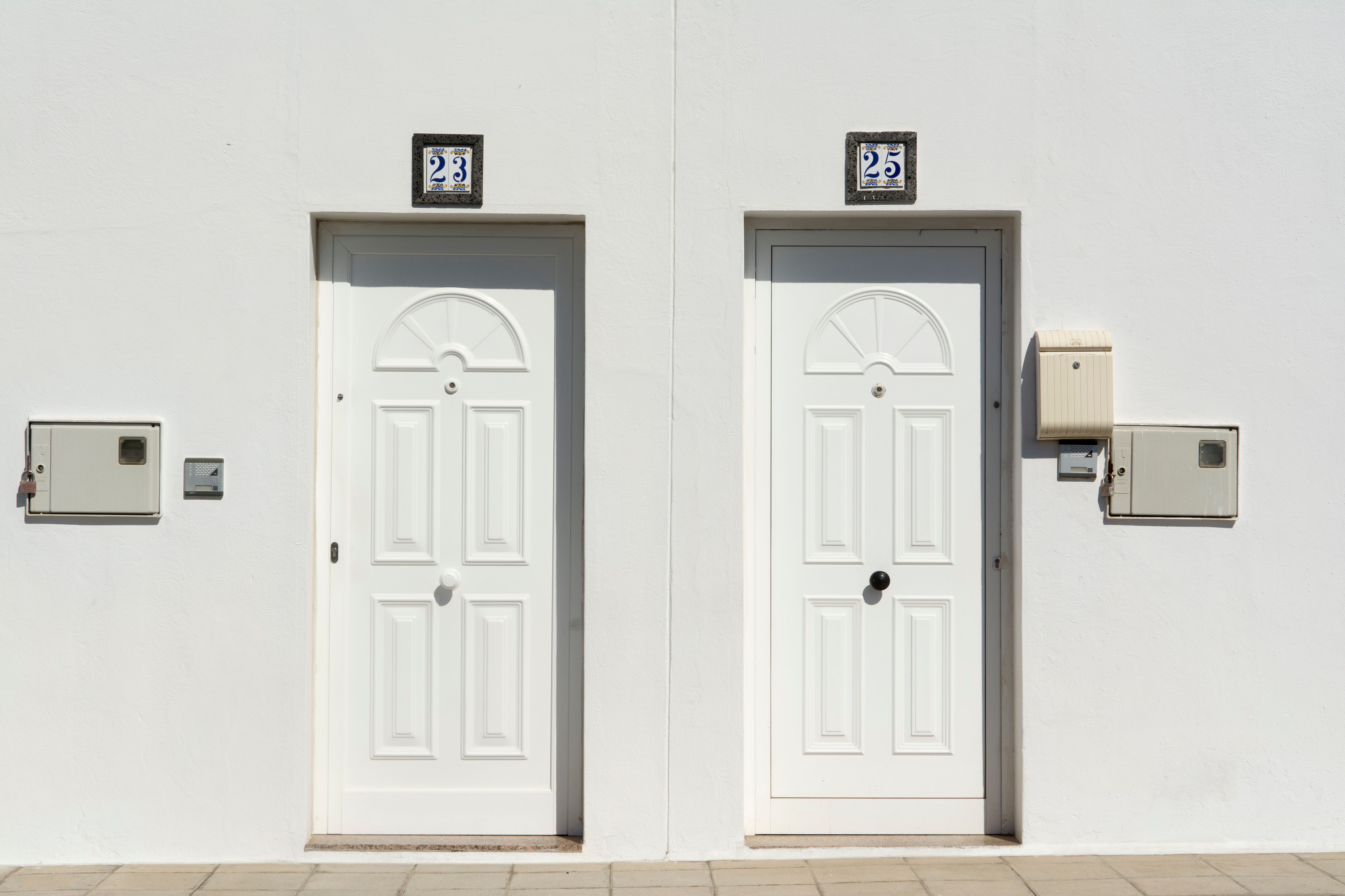 Duas portas brancas idênticas, lado a lado, em uma parede branca. Acima da porta esquerda, um azulejo com o número 23. Acima da porta direita, um azulejo com o número 25. Ambas as portas têm maçanetas redondas e painéis decorativos. À esquerda da porta 23, há uma caixa de medidor e um interfone. À direita da porta 25, há uma caixa de correio e uma caixa de medidor. O chão é de lajotas claras. A luz do sol cria sombras nítidas