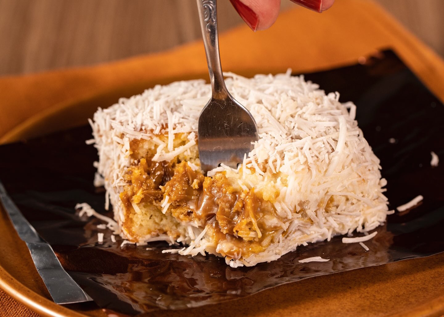 Um pedaço de bolo branco coberto com coco ralado, recheado com doce de leite, sendo cortado por um garfo. O bolo está sobre um prato marrom escuro com uma folha de papel manteiga preta, em um fundo de madeira e tecido laranja