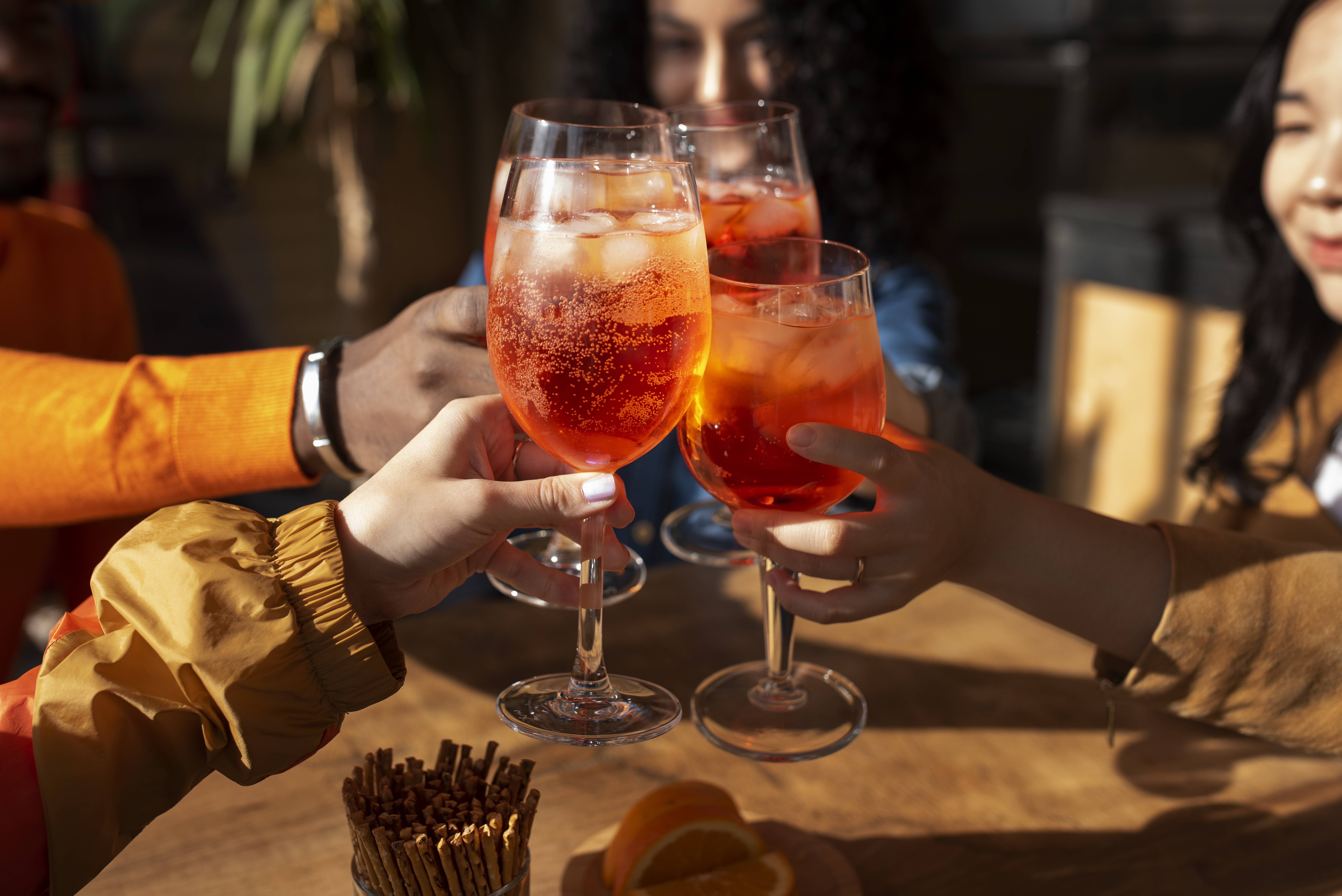 Quatro pessoas brindando com ta&ccedil;as de Aperol Spritz, bebidas alaranjadas com gelo e bolhas. As m&atilde;os seguram as ta&ccedil;as no centro da imagem, com um homem de blusa laranja &agrave; esquerda e duas mulheres sorrindo ao fundo. Uma mesa de madeira com biscoitos salgados e fatias de laranja aparece em primeiro plano