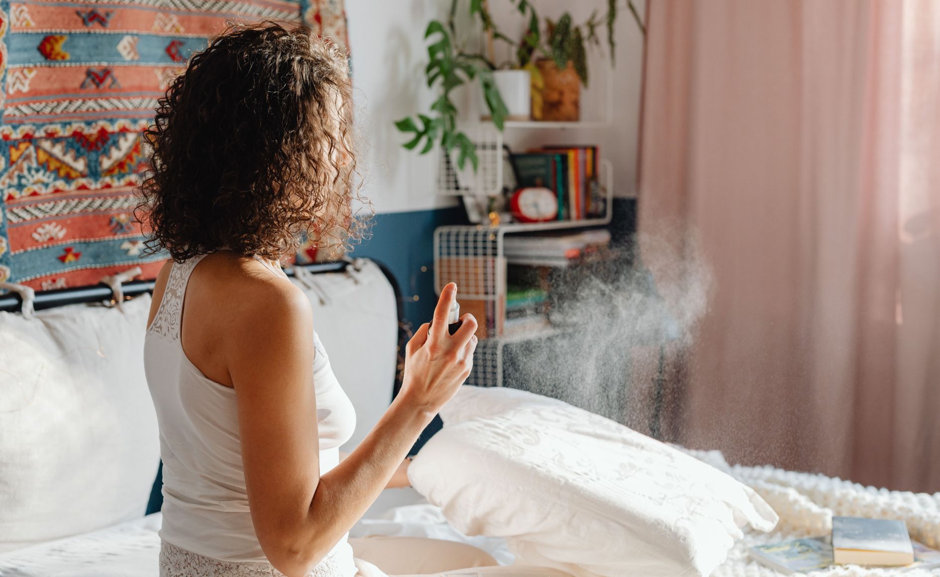 Mulher de cabelos cacheados, vista de costas, borrifa spray em um travesseiro branco sobre a cama. Ela veste regata branca e está em um quarto com tapete colorido na parede e prateleiras com livros