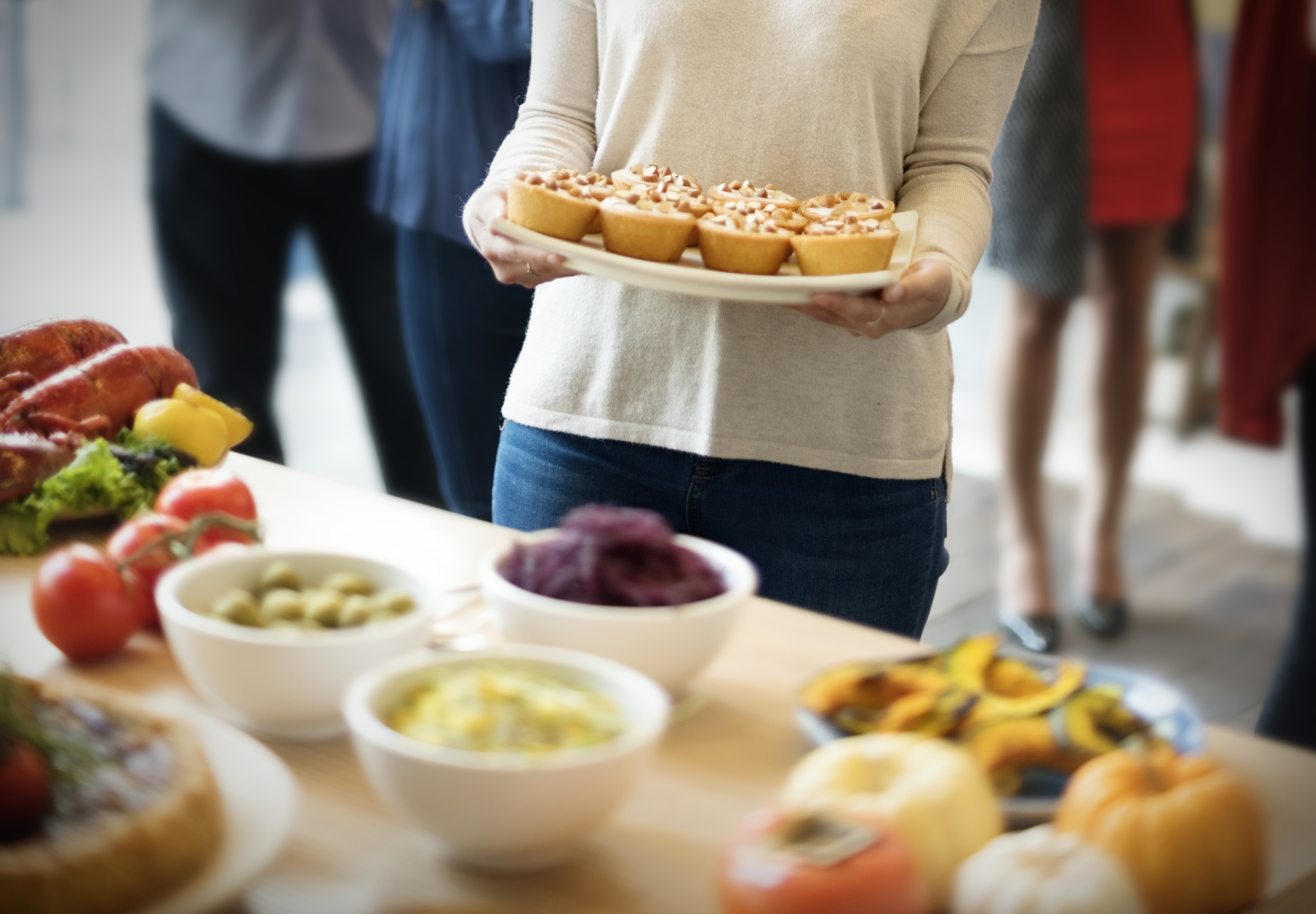 Pessoa de suéter bege e jeans segura bandeja com seis tortinhas doces, enquanto outras pessoas desfocadas e uma mesa com diversos alimentos, como lagosta, tomates, azeitonas e abóbora, preenchem o primeiro plano e o fundo