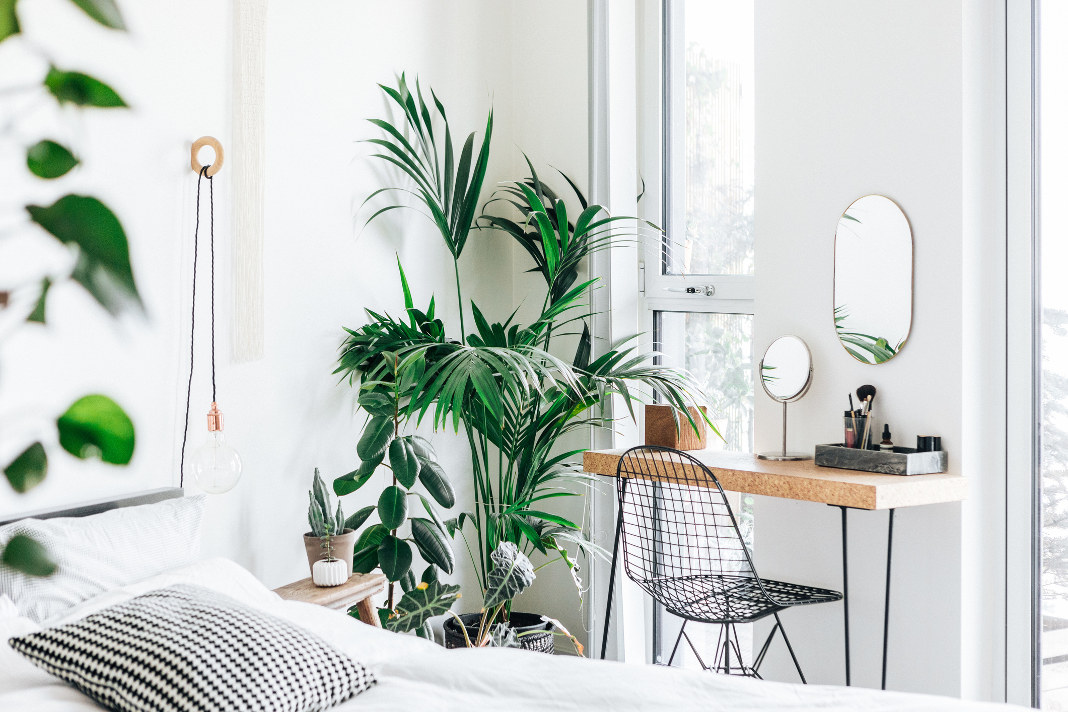 Quarto claro com cama branca, almofada xadrez, plantas verdes, mesa de cabeceira de madeira, penteadeira com espelhos e cadeira de metal preta, ao lado de uma janela grande