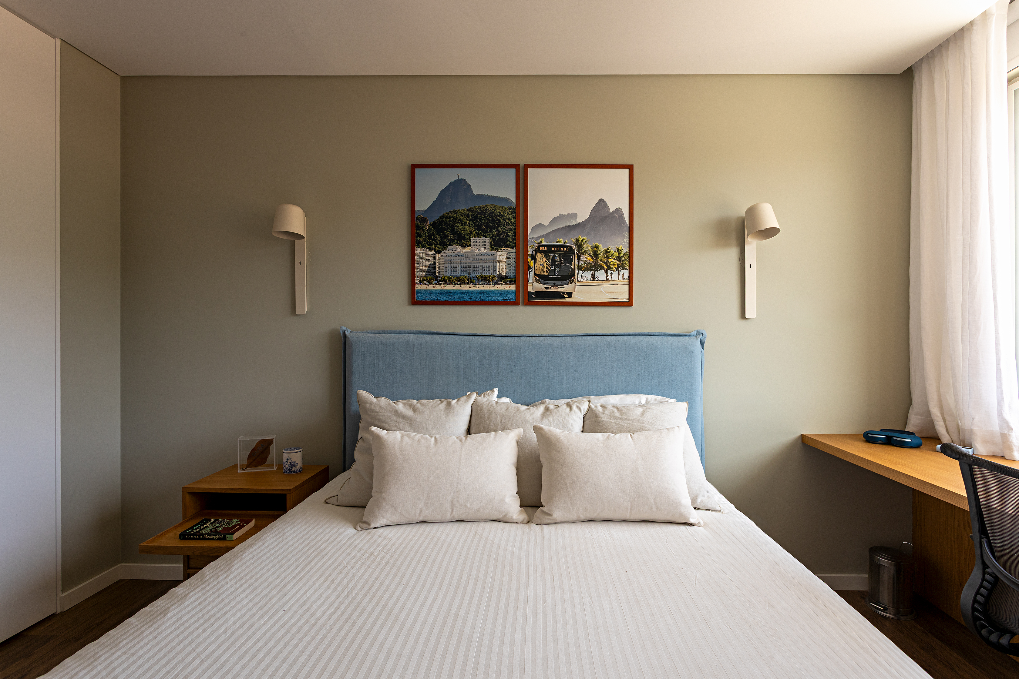 Quarto moderno com cama de casal, cabeceira azul, len&ccedil;&oacute;is brancos listrados e travesseiros claros. Na parede verde-clara, duas arandelas brancas e dois quadros com paisagens do Rio de Janeiro. &Agrave; esquerda, uma mesa de cabeceira de madeira com livro e objeto decorativo. &Agrave; direita, uma escrivaninha de madeira clara com cadeira preta e cortina branca na janela