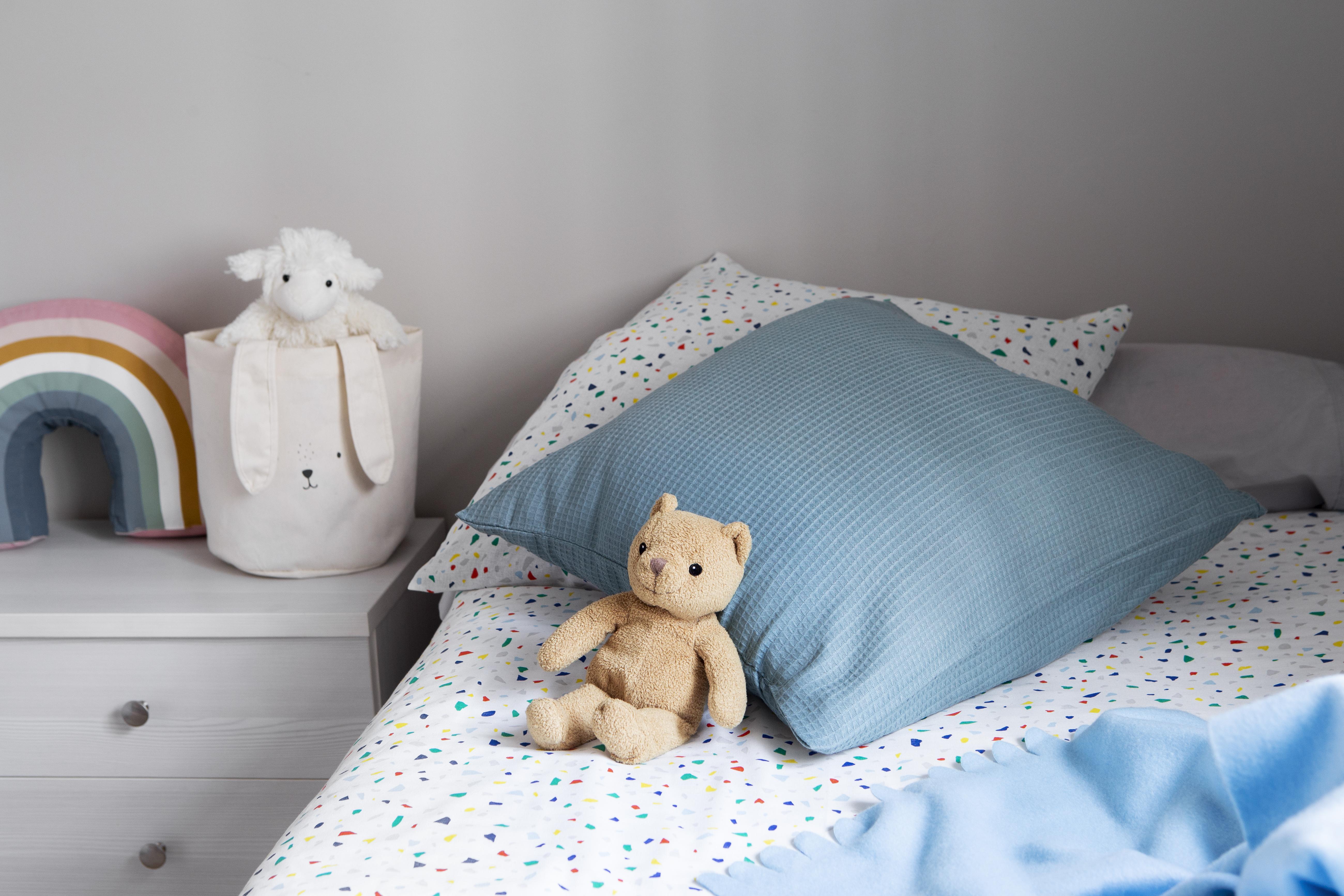 Quarto infantil com cama arrumada, lençol branco de confetes coloridos, travesseiro azul texturizado e um ursinho de pelúcia marrom sentado. Na mesa de cabeceira branca, um cesto de tecido com orelhas de coelho e um carneiro de pelúcia, ao lado de uma almofada de arco-íris