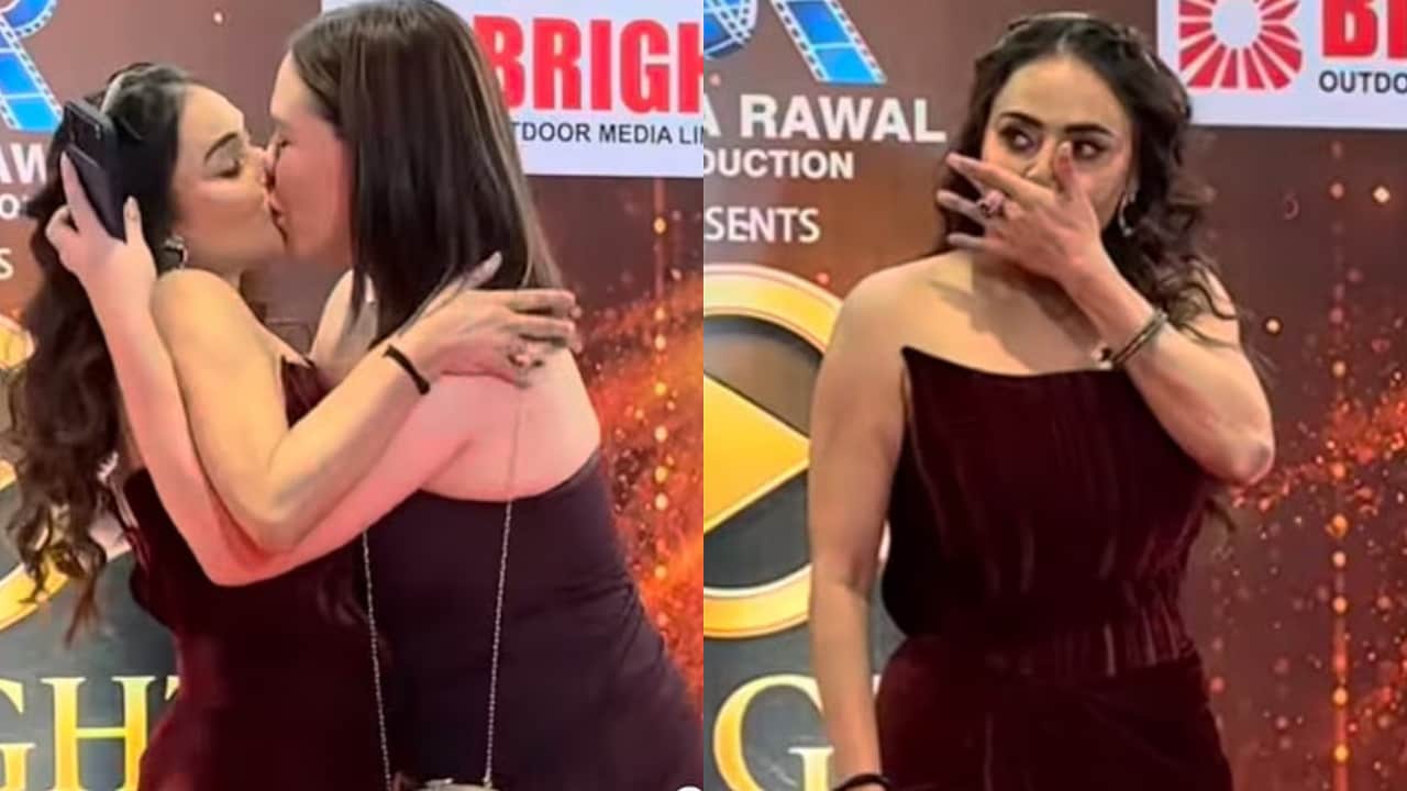 Duas mulheres em um evento. À esquerda, uma mulher de cabelo escuro e vestido vinho beija outra mulher de cabelo escuro. À direita, a mulher de vestido vinho cobre a boca com a mão, parecendo surpresa ou envergonhada. O fundo tem logos de empresas