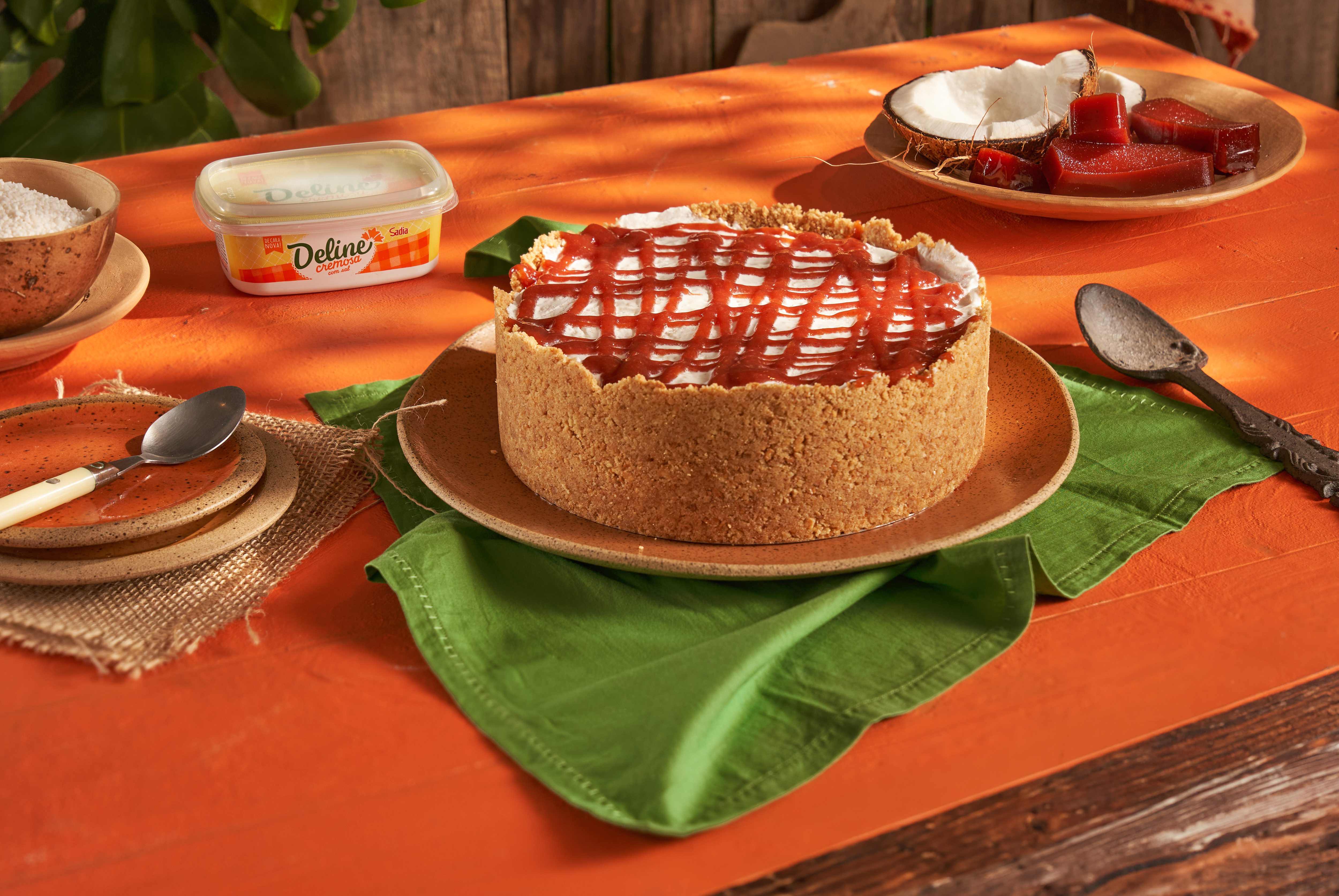 Cheesecake com base de biscoito e cobertura branca, decorado com calda vermelha em zigue-zague, sobre prato marrom e guardanapo verde. Ao fundo, uma embalagem de margarina Del&iacute;cia, uma tigela com coco ralado, e outra com peda&ccedil;os de goiabada e coco fresco. Tudo disposto sobre uma mesa de madeira com um caminho de mesa laranja
