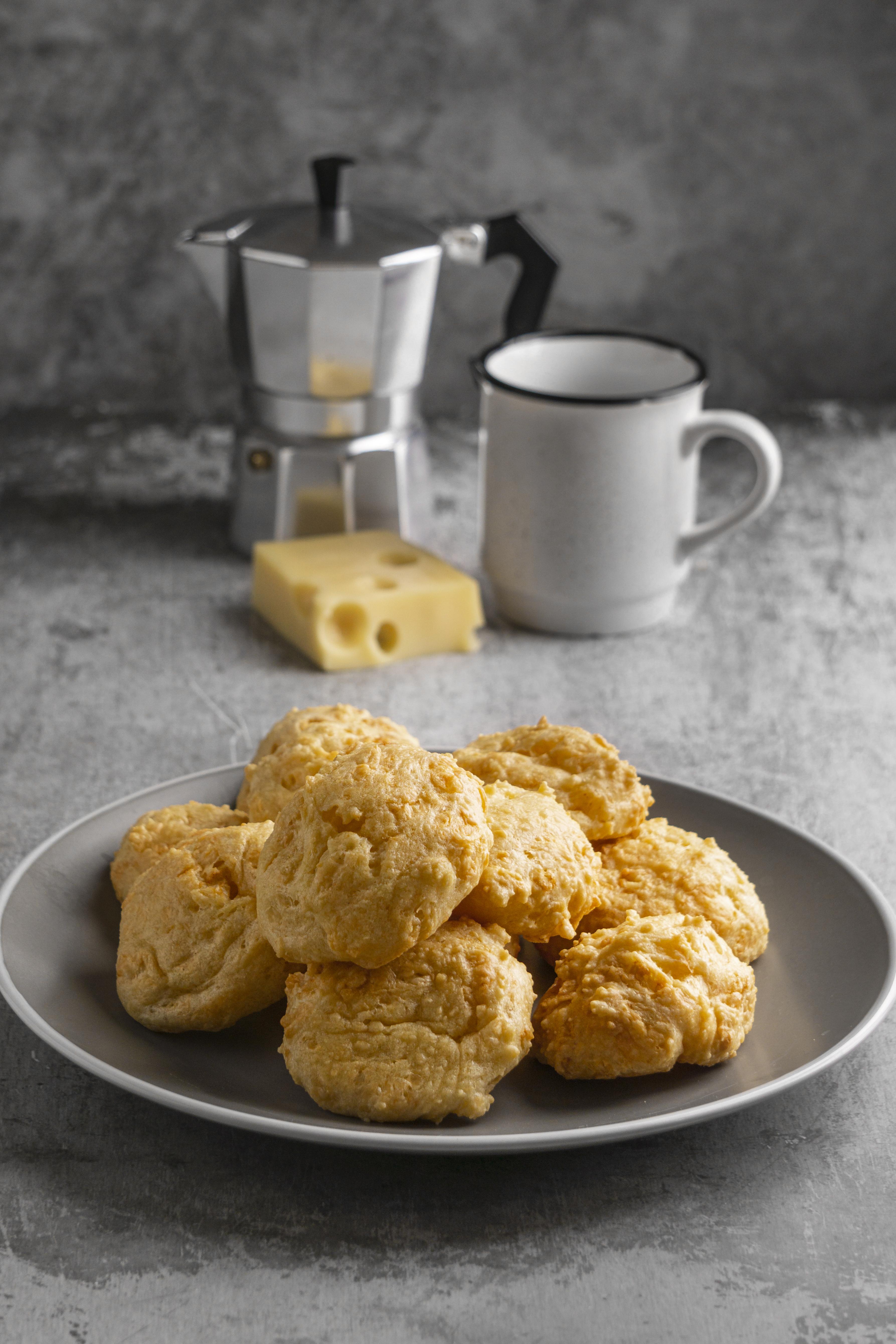 P&atilde;es de queijo dourados em um prato cinza, com uma cafeteira italiana, uma caneca branca e um peda&ccedil;o de queijo su&iacute;&ccedil;o ao fundo, sobre uma superf&iacute;cie de cimento cinza