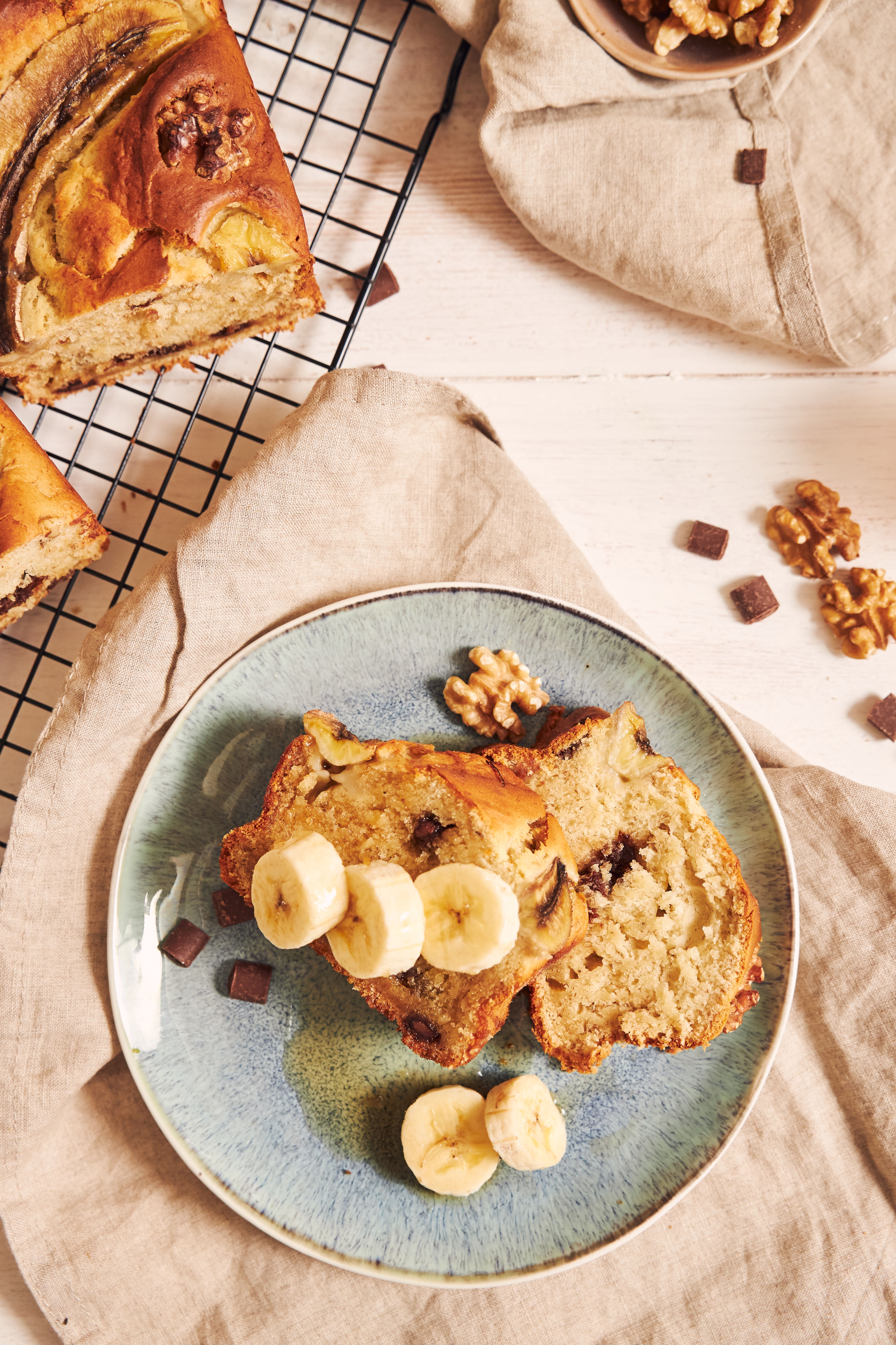 Fatias de bolo de banana com gotas de chocolate e nozes, decoradas com rodelas de banana e nozes, sobre um prato azul e uma toalha de linho em uma mesa branca