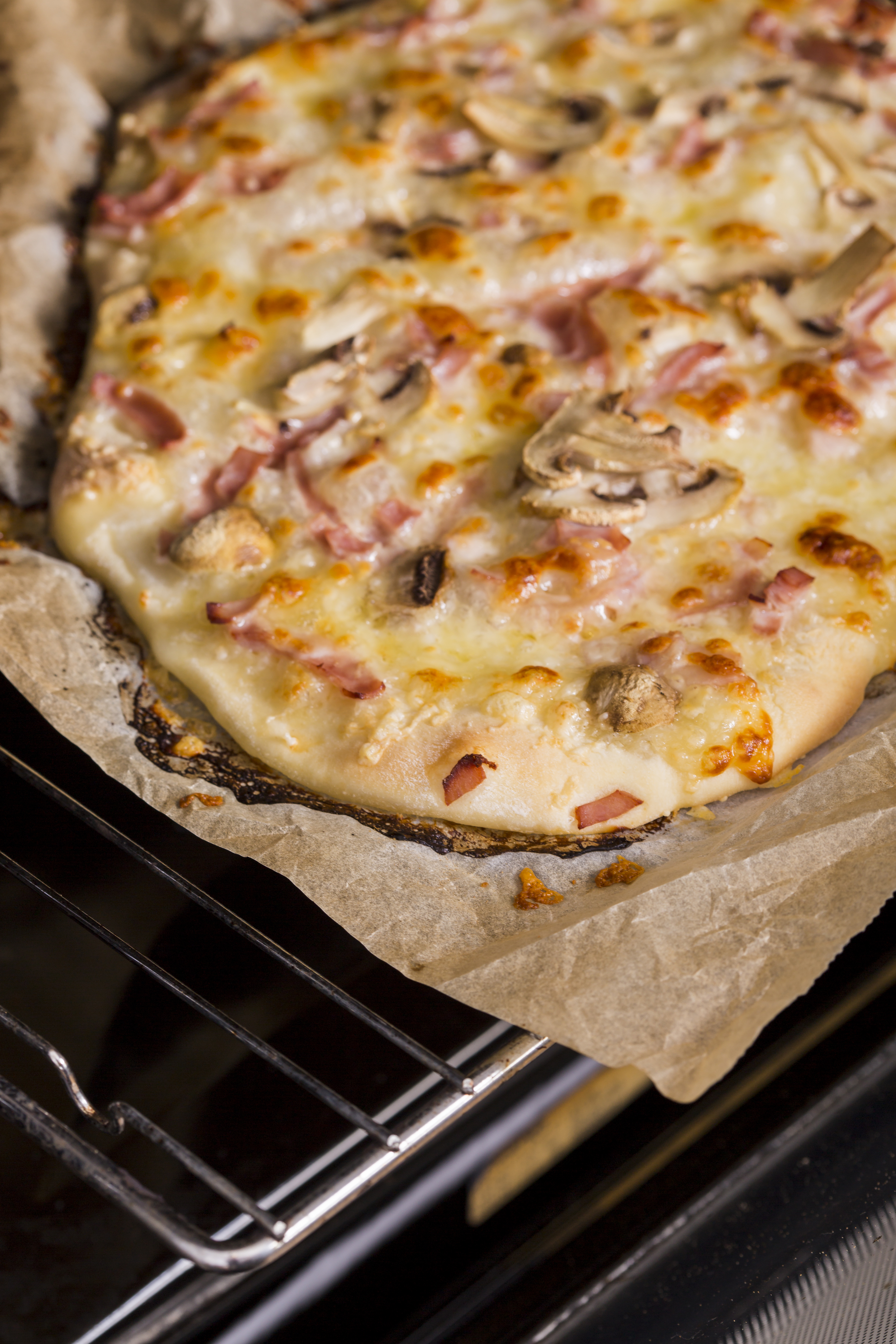 Pizza de massa fina com queijo derretido, presunto e cogumelos, assando em papel manteiga sobre uma grade de forno
