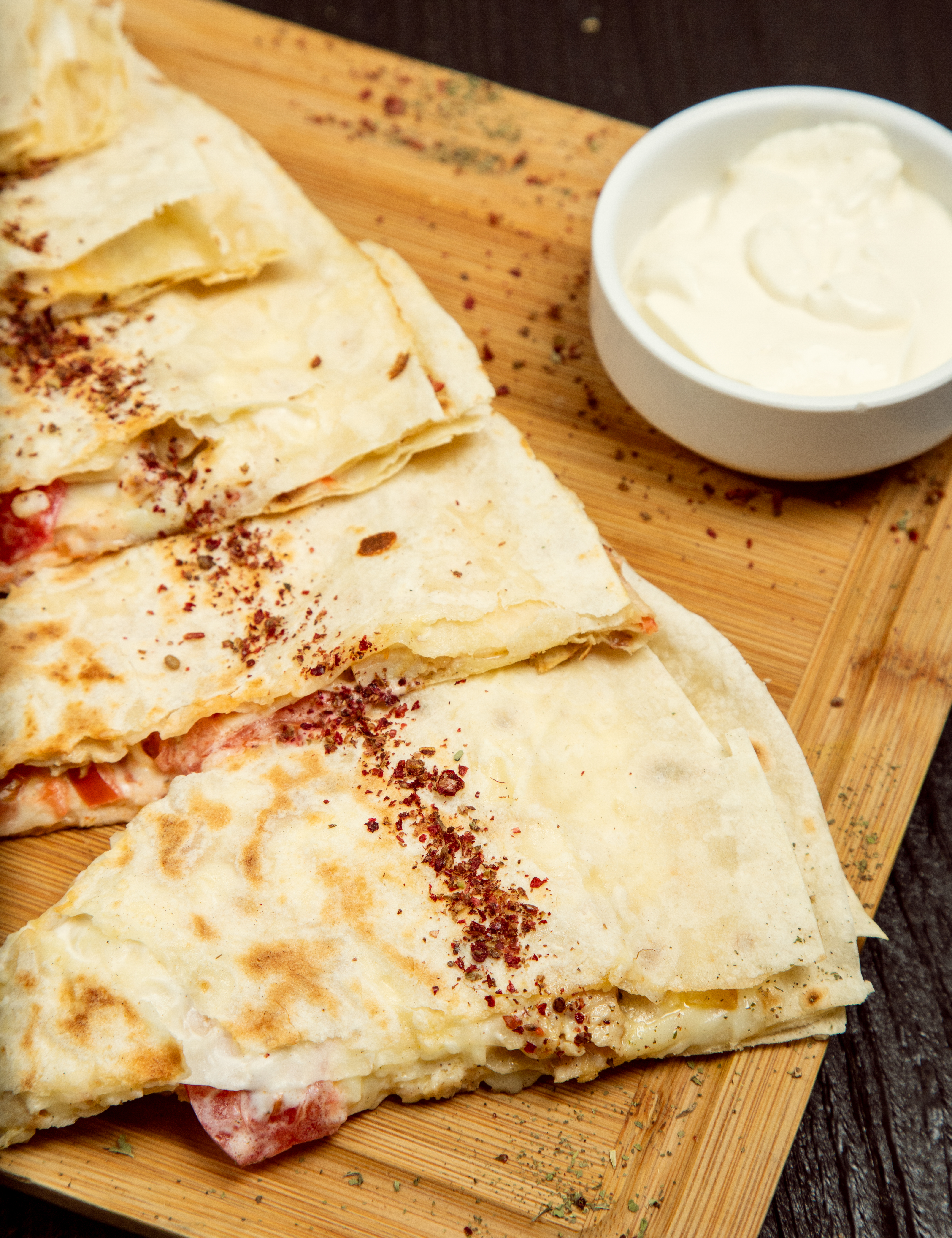 Tr&ecirc;s fatias de quesadilla recheadas com queijo derretido e tomate, polvilhadas com especiarias vermelhas e verdes, servidas em uma t&aacute;bua de madeira clara ao lado de um pote branco com molho cremoso