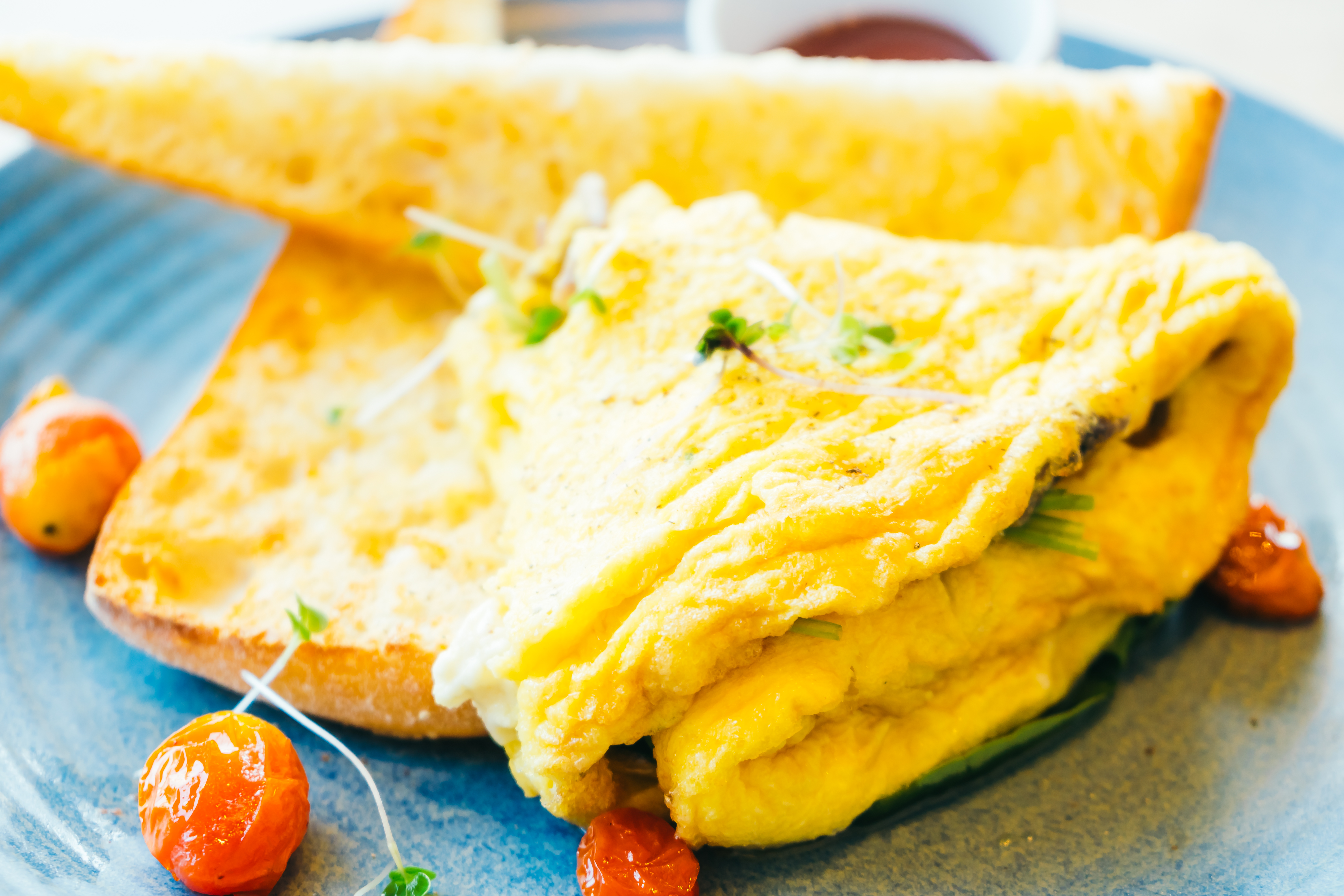 Omelete amarelo dobrado com brotos verdes, acompanhado de torradas douradas e tomates cereja em um prato azul