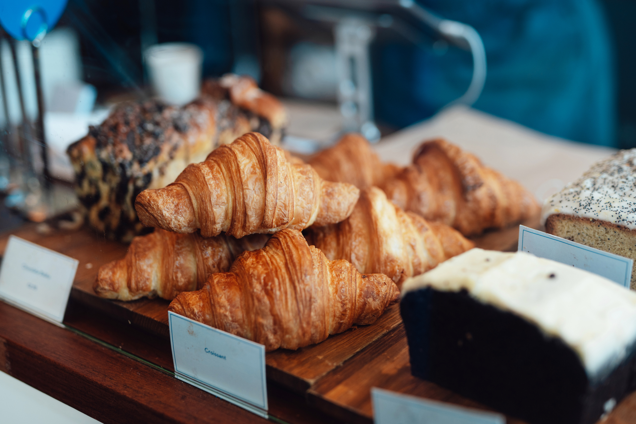 Pães e doces em uma vitrine de padaria, com croissants dourados em primeiro plano, um bolo escuro com cobertura branca e um pão com sementes de papoula ao fundo. Placas de identificação com nomes dos produtos estão visíveis na frente