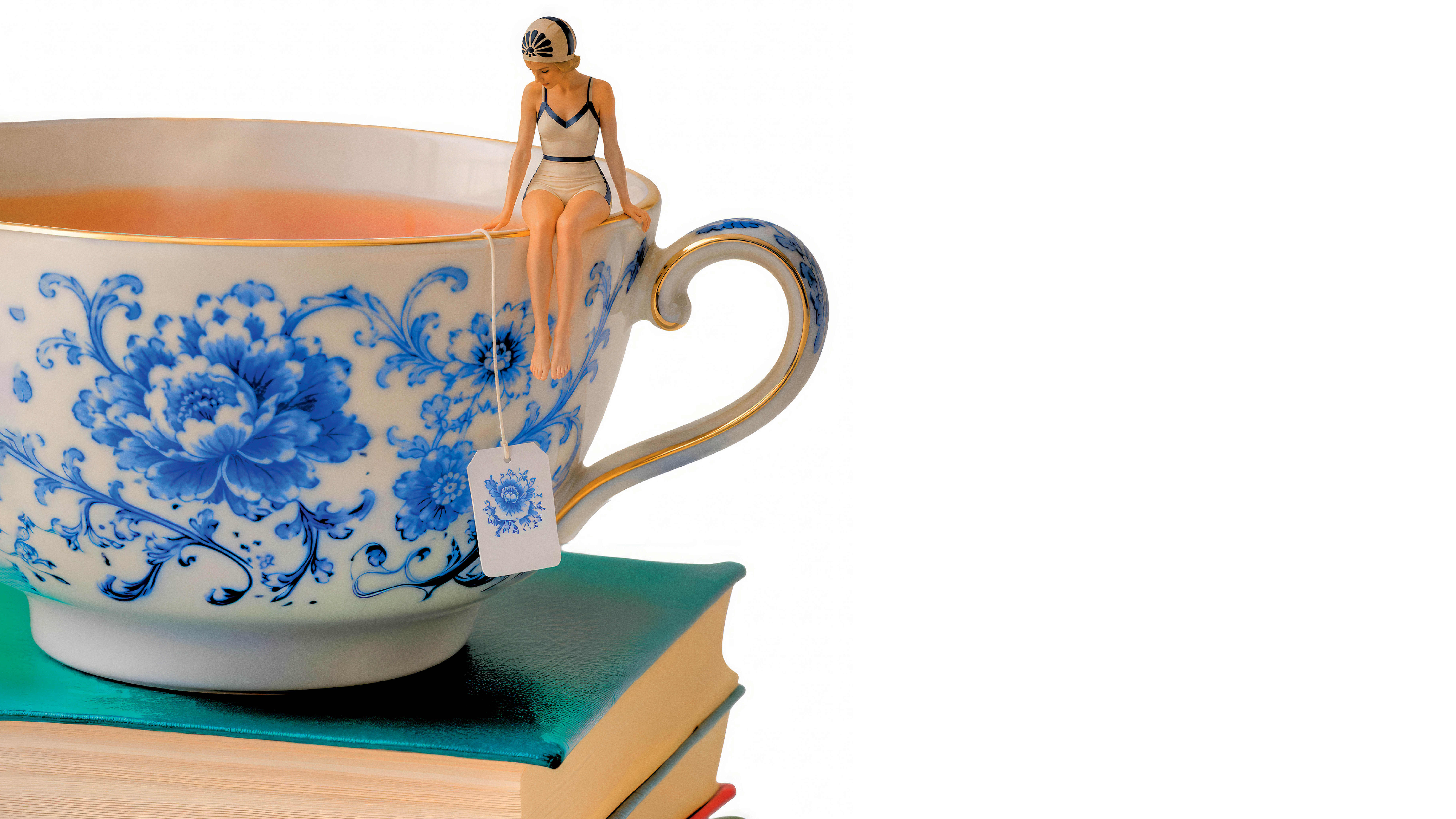 Uma miniatura de mulher em traje de banho vintage sentada na borda de uma xícara de chá grande, decorada com flores azuis e dourado, com um saquinho de chá pendurado na mão. A xícara está sobre uma pilha de livros com capas verdes, em um fundo branco