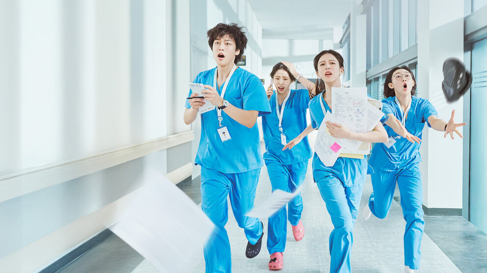 Quatro profissionais de sa&uacute;de asi&aacute;ticos, vestindo uniformes azuis, correm apressadamente por um corredor de hospital, com express&otilde;es de surpresa e urg&ecirc;ncia. Pap&eacute;is voam ao redor deles, e um sapato preto &eacute; arremessado no ar &agrave; direita, indicando a pressa.