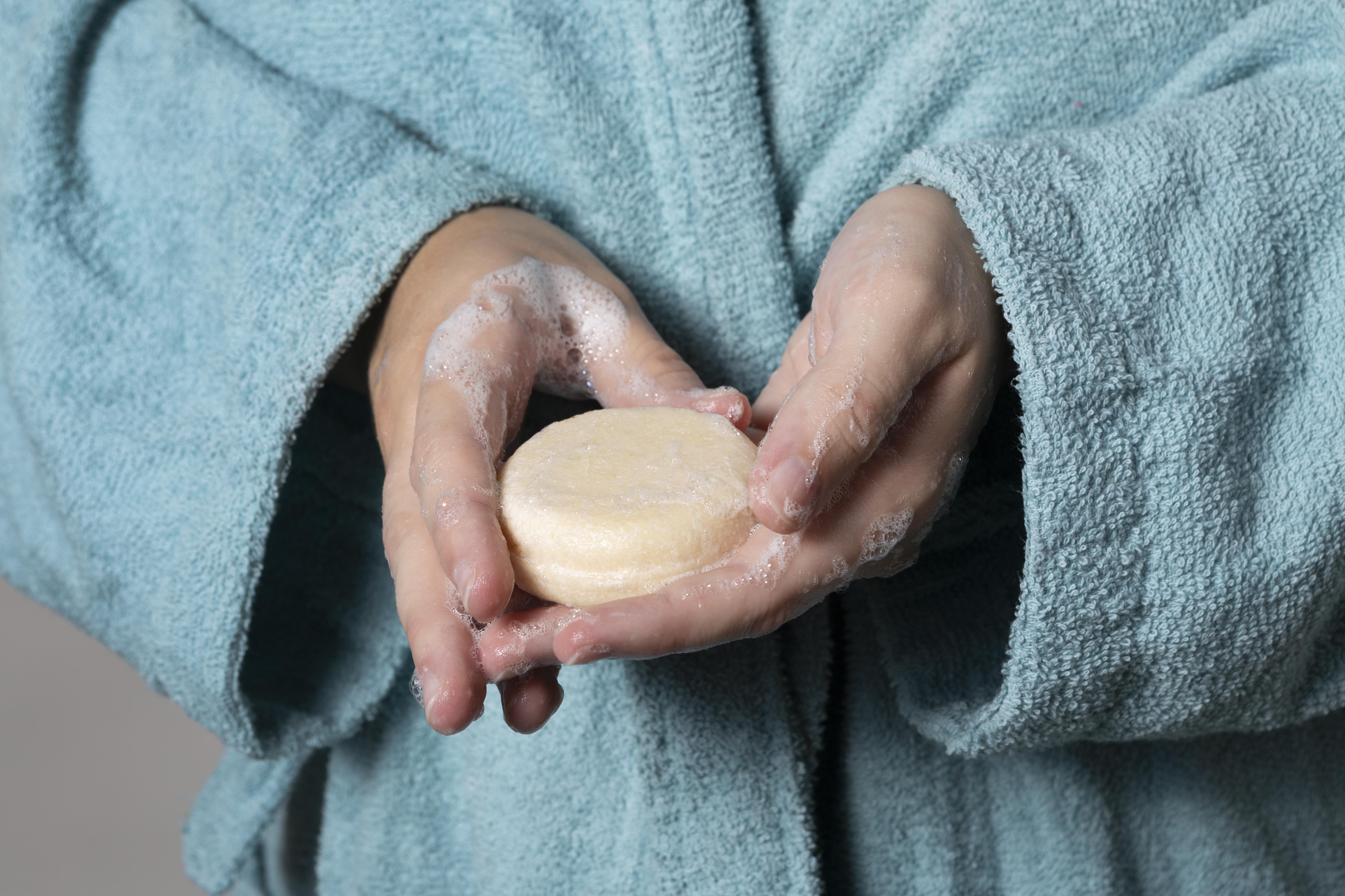 Mãos de uma pessoa vestindo um roupão azul-turquesa segurando um sabonete redondo bege com espuma branca nas palmas e dedos