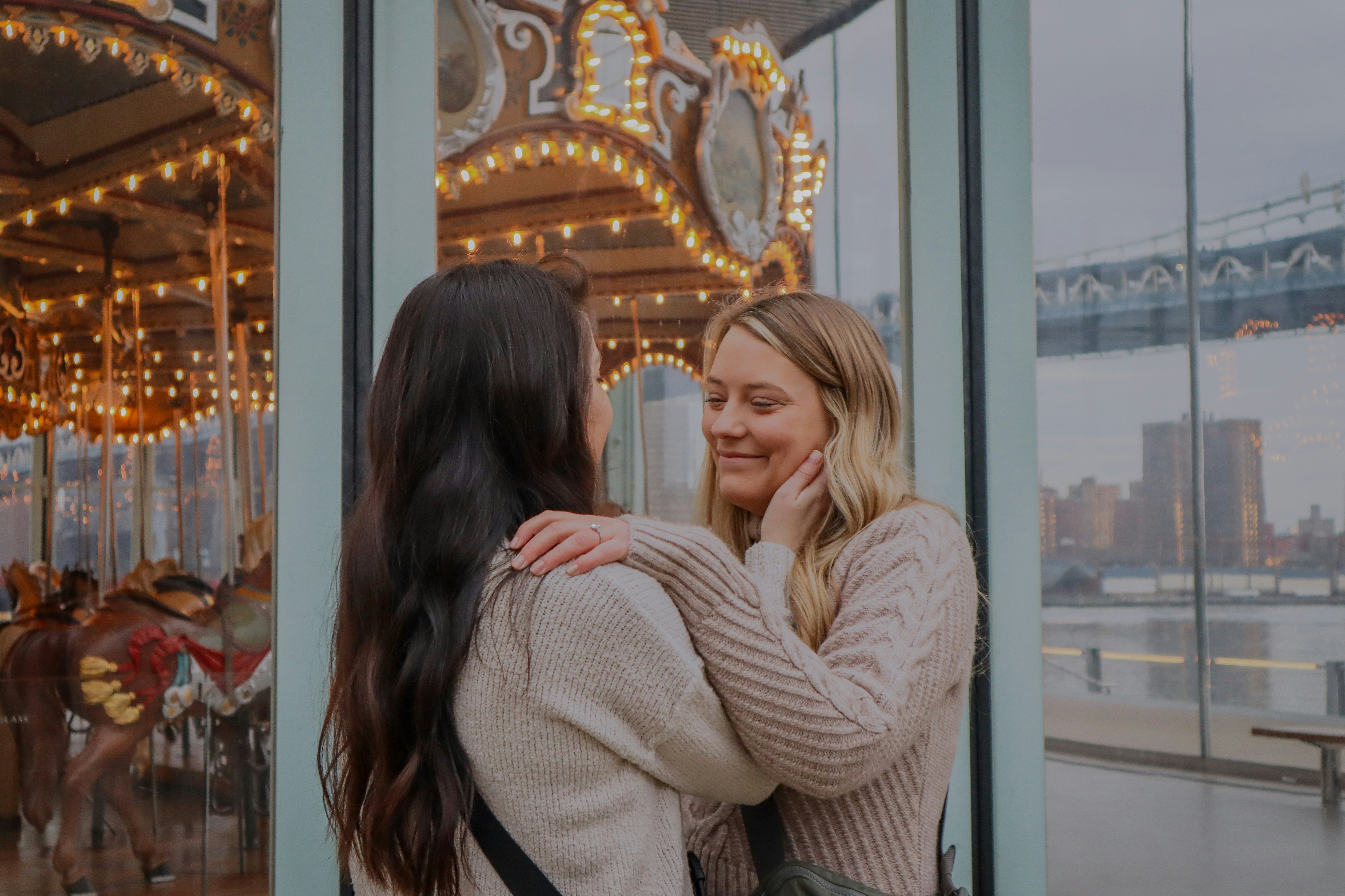 Duas mulheres, uma loira e uma morena, abra&ccedil;adas e sorrindo, com um carrossel iluminado e uma ponte ao fundo, atrav&eacute;s de uma janela de vidro