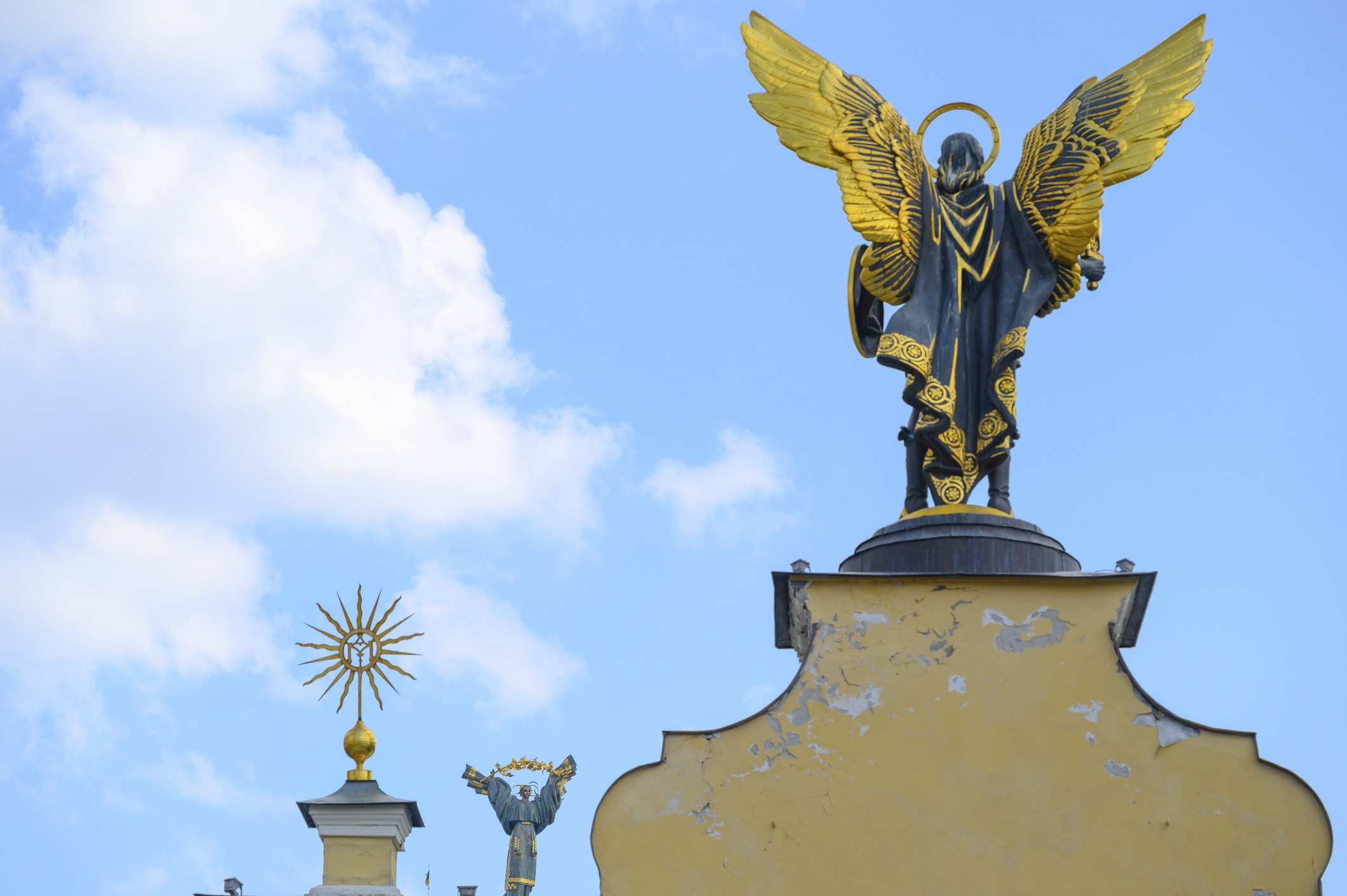 Estátua de um anjo com asas douradas e manto escuro, visto de costas, sobre um pedestal amarelo desgastado, sob céu azul com nuvens brancas. À esquerda, um ornamento dourado em forma de sol.
