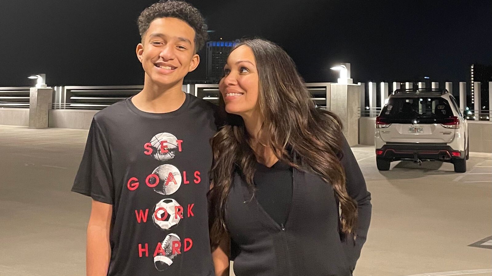 Um adolescente sorridente com cabelo cacheado e uma mulher de cabelos longos e castanhos, ambos vestindo preto, em um estacionamento noturno. O garoto usa uma camiseta com SET GOALS WORK HARD e imagens de bolas esportivas. A mulher olha para ele com carinho. Um carro branco est&aacute; estacionado ao fundo