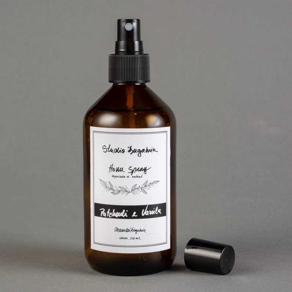 Frasco de vidro &acirc;mbar com borrifador preto, contendo Home Spray Patchouli e Vanila da Studio Bagamin, 250 ml, sobre fundo cinza. A tampa do borrifador est&aacute; ao lado