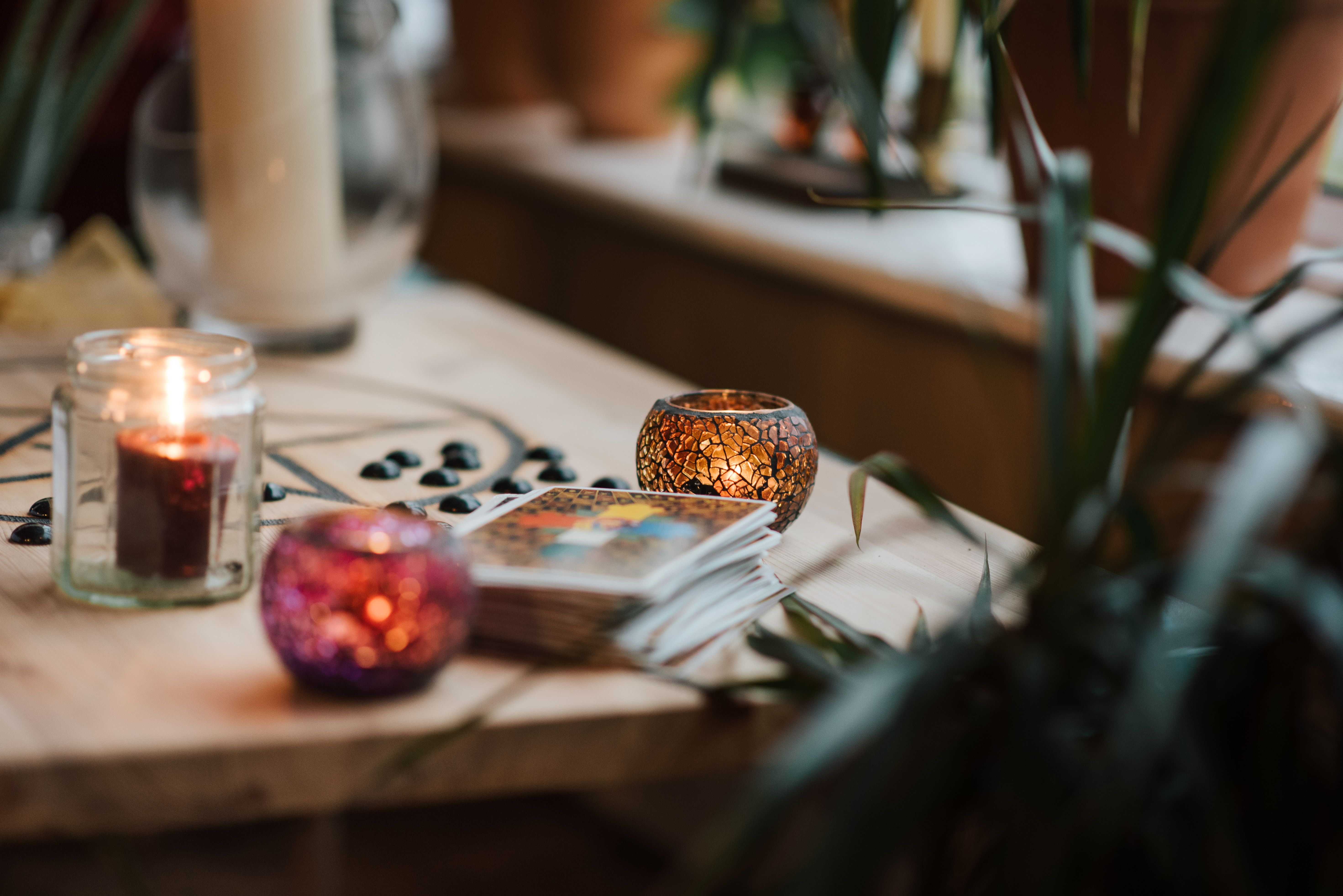 Uma mesa de madeira clara com uma vela acesa em pote de vidro, um castiçal de mosaico laranja e outro roxo, um baralho de cartas de tarô e pedras pretas. Ao fundo, plantas em vasos e uma janela, criando uma atmosfera mística