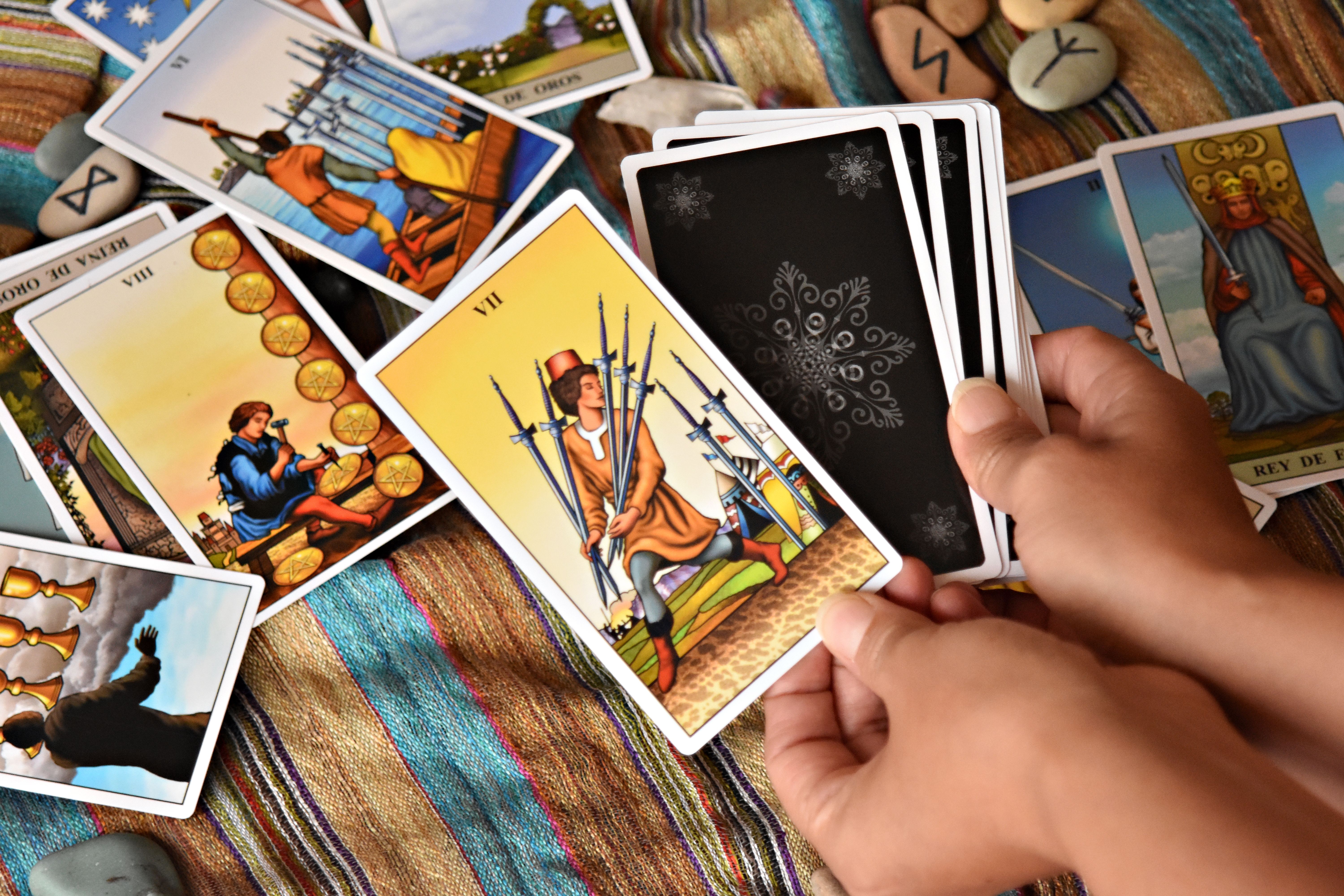 Mãos seguram cartas de tarô com o verso preto e desenhos brancos, enquanto outras cartas coloridas estão espalhadas sobre um tecido listrado. Uma carta central mostra um homem com sete espadas.