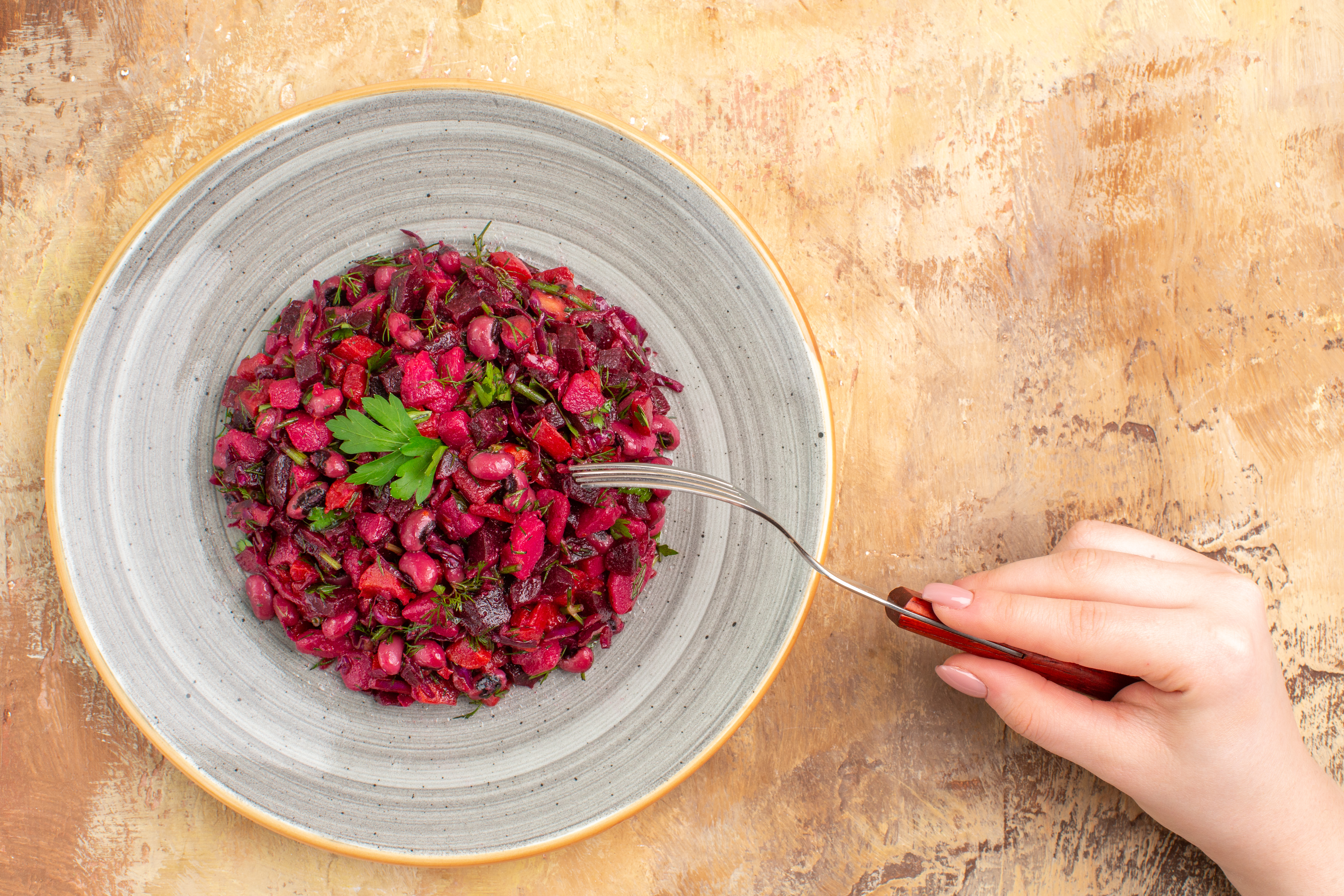 Salada vibrante de beterraba e feij&atilde;o vermelho em prato cinza, com garfo sendo segurado por m&atilde;o feminina &agrave; direita, sobre fundo texturizado marrom claro