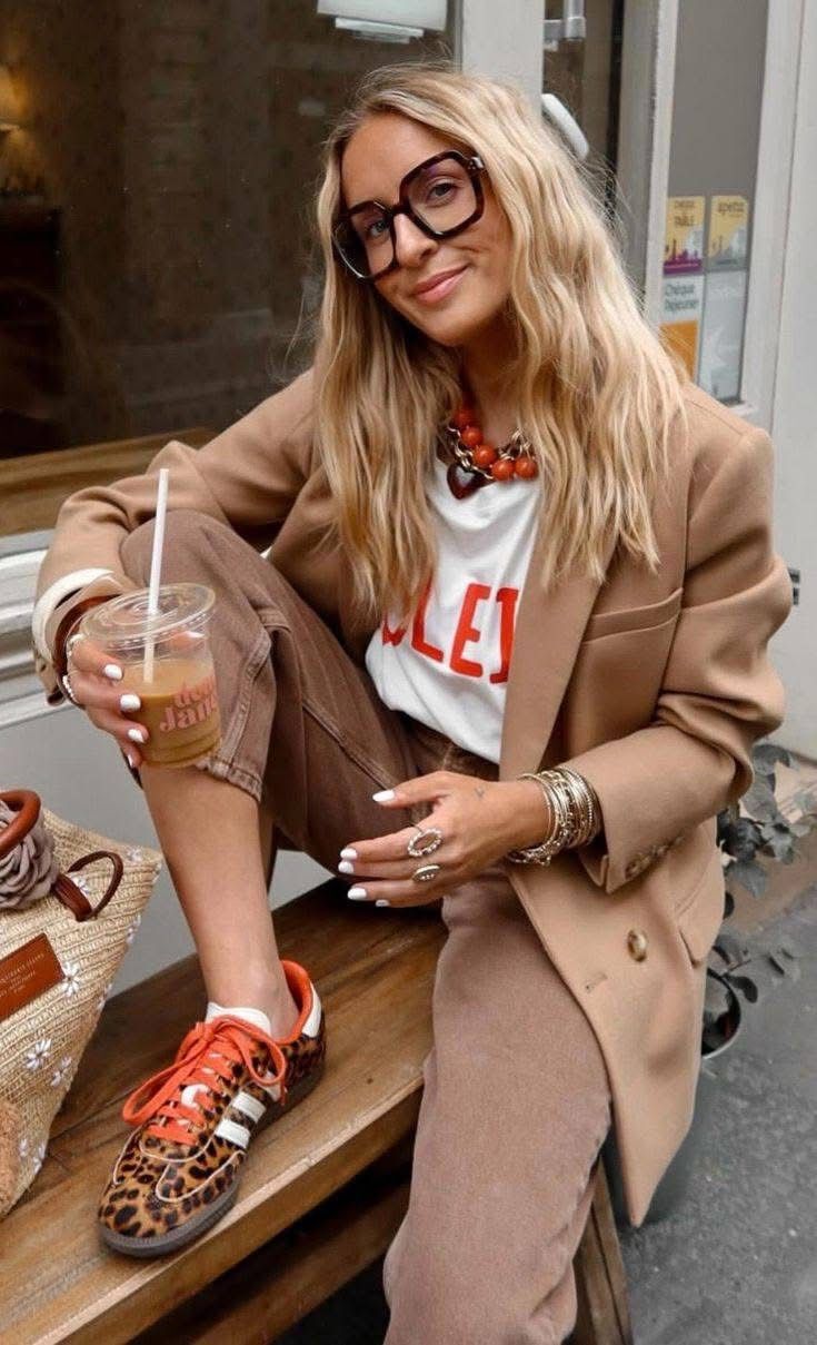 Mulher loira sorridente, com &oacute;culos grandes e blazer bege, sentada em um banco de madeira, segurando um copo de caf&eacute; gelado. Ela usa camiseta branca com letras vermelhas, cal&ccedil;a marrom, t&ecirc;nis de oncinha com cadar&ccedil;os laranja e an&eacute;is e pulseiras douradas. Ao fundo, uma vitrine com folhetos