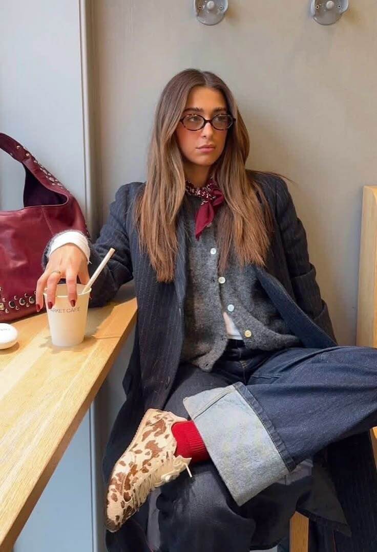 Mulher jovem com cabelo castanho e &oacute;culos ovais sentada em um caf&eacute;, vestindo blazer listrado, cardig&atilde; cinza, cal&ccedil;a jeans larga com barra dobrada, meias vermelhas e t&ecirc;nis com estampa de vaca. Ela segura um copo de papel com canudo e uma bolsa vermelha est&aacute; ao seu lado