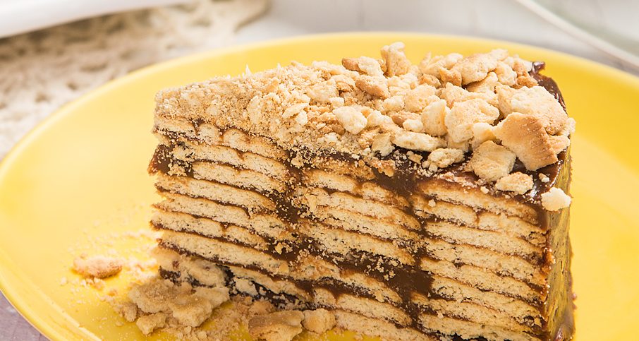 Fatia de torta de bolacha com camadas de creme de chocolate e cobertura de farofa de biscoito, servida em prato amarelo sobre mesa branca com toalha de renda